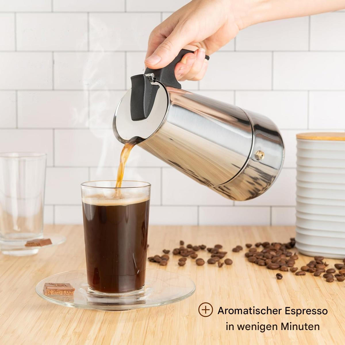 Eine Hand gießt Kaffee aus einer Silberkanne in ein Glas mit dunklem Kaffee. Kaffeebohnen liegen auf dem Tisch.