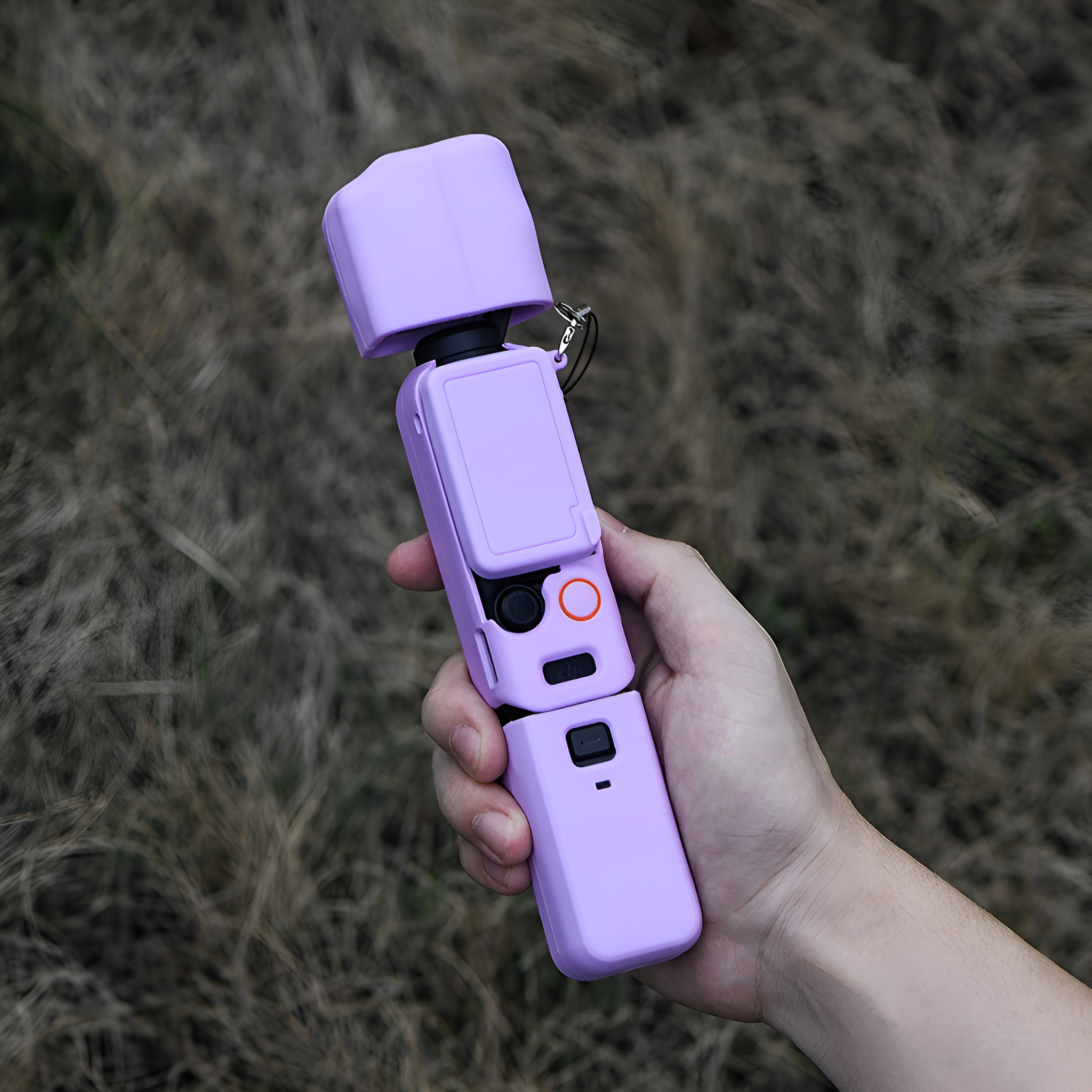 Eine lila Handkamera mit Hülle wird in einer Hand gehalten, mit verschwommenem Hintergrund aus trockenem Gras.