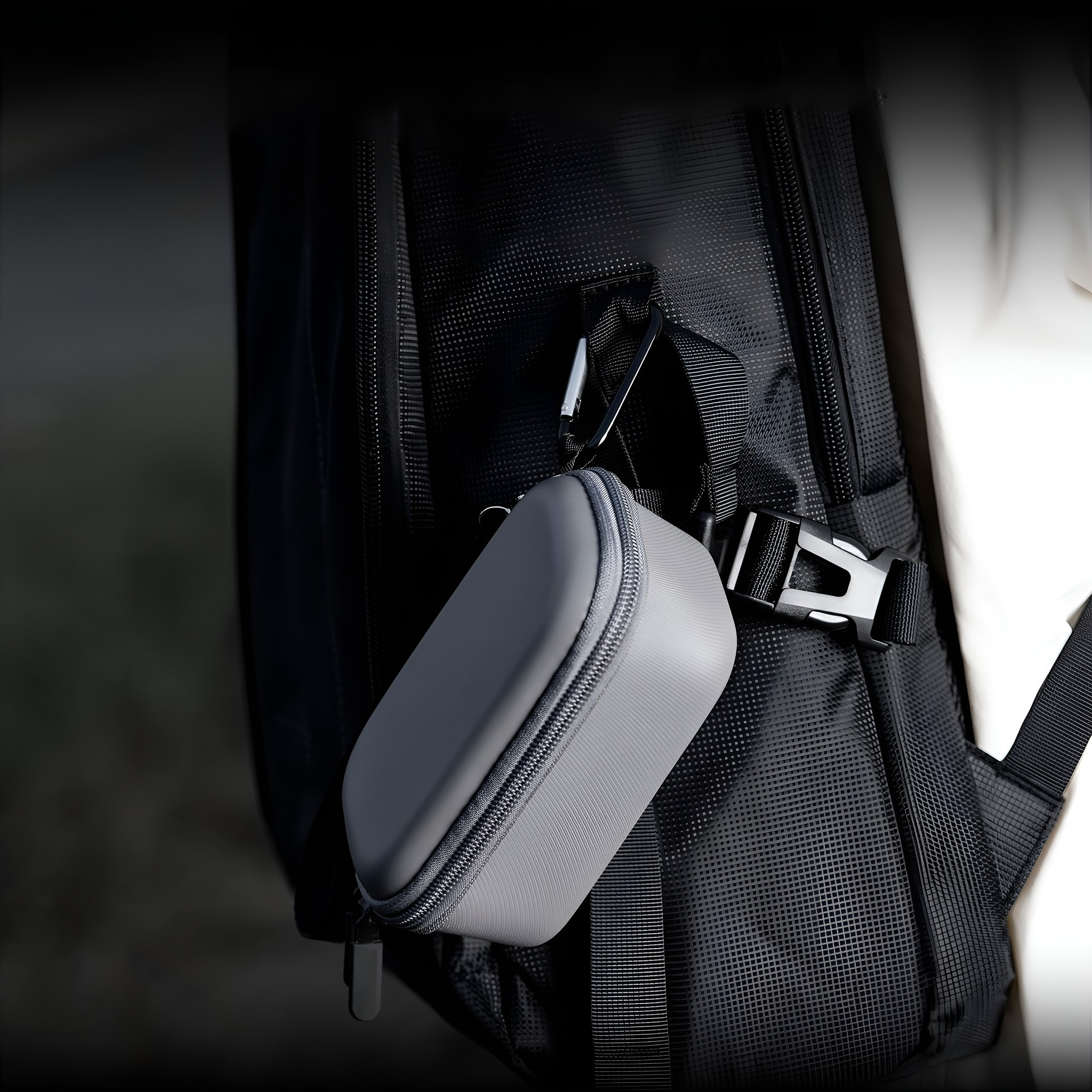 Ein graues Etui, das mit einem silbernen Karabinerhaken an einem schwarzen Rucksack befestigt ist.
