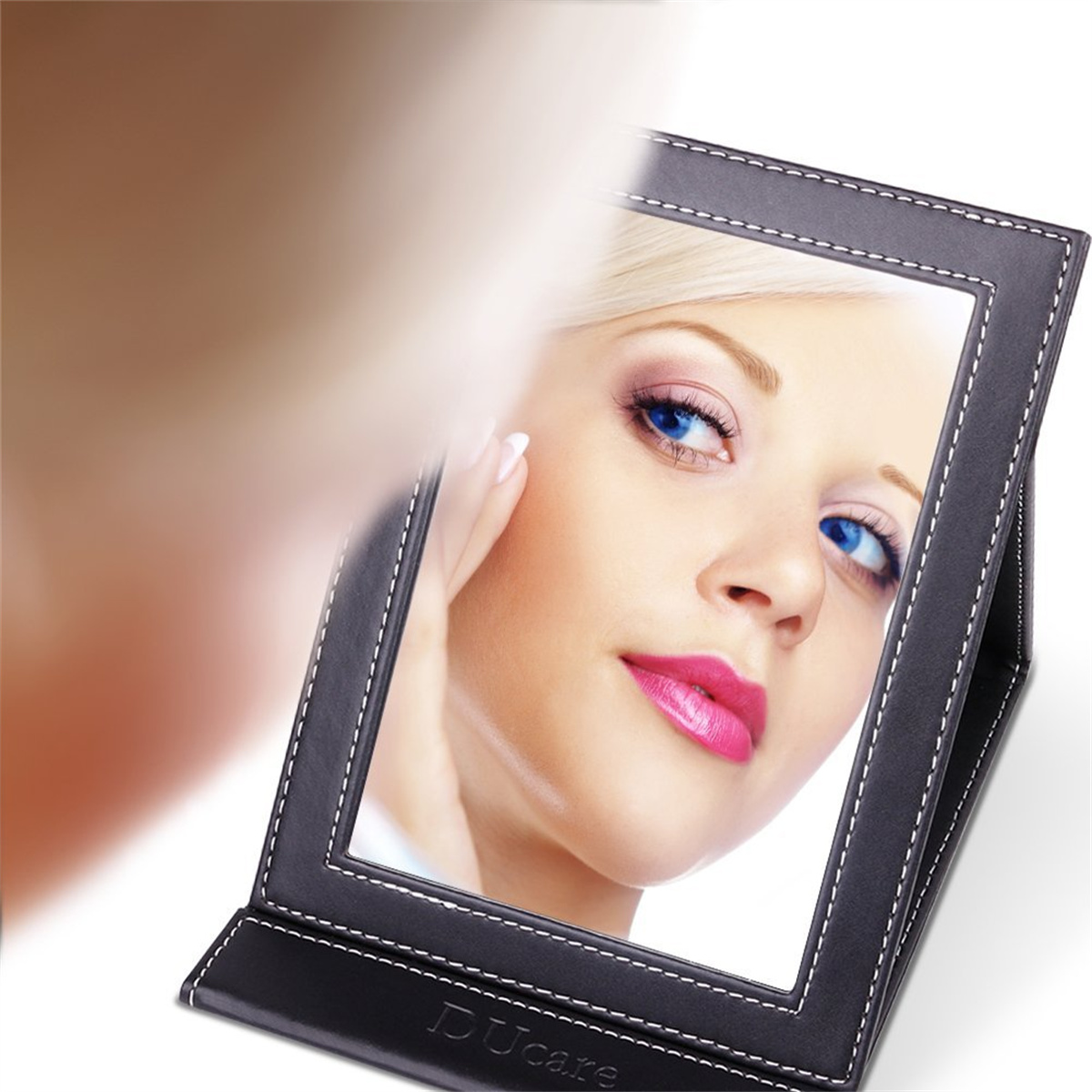 Eine Frau blickt in einen schwarzen Spiegel, Nahaufnahme ihres Gesichts. Sie trägt rosa Lippenstift und hat blaue Augen.