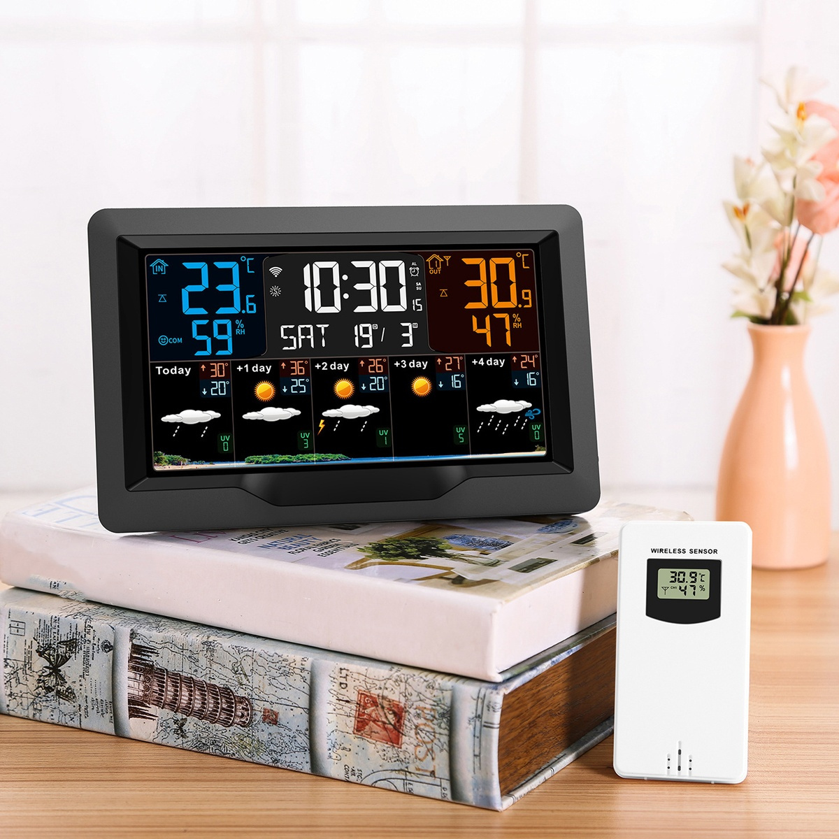 Eine schwarze Wetterstation zeigt Zeit, Temperatur und Vorhersage. Ein weißer Sensor liegt auf einem Buch.