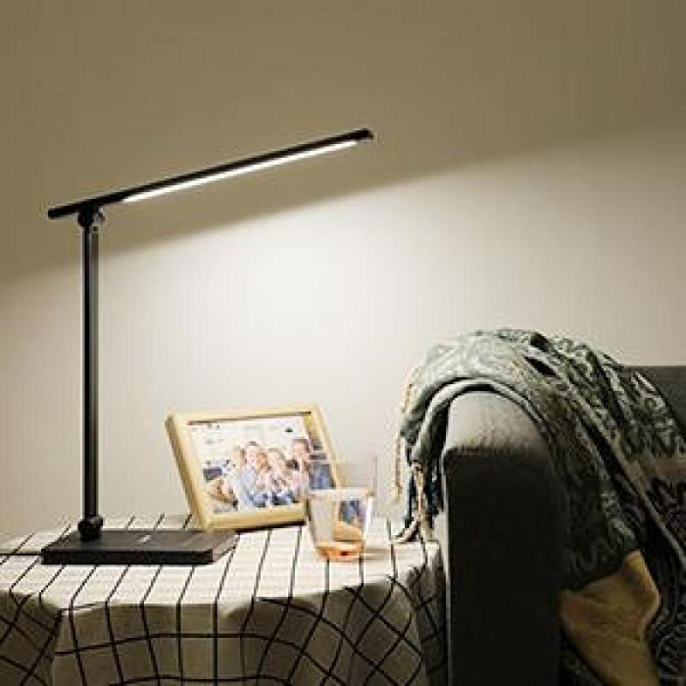Eine schwarze Schreibtischlampe beleuchtet einen Tisch mit einem gerahmten Foto, einem Glas und einer karierten Tischdecke; Hintergrund ist eine Couch.