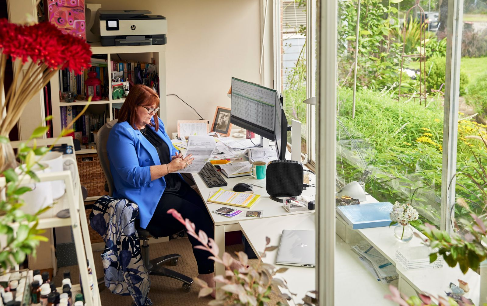 Une femme en blazer bleu examine des documents à un bureau. Des étagères et une imprimante sont en arrière-plan. Dehors, une fenêtre, de la verdure.