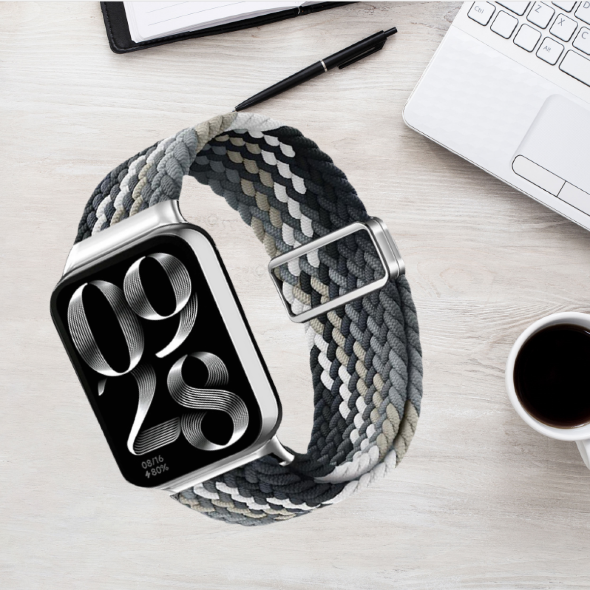 Smartwatch mit mehrfarbig geflochtenem Armband auf einem Holzschreibtisch mit Stift, Notizblock und Kaffeetasse.