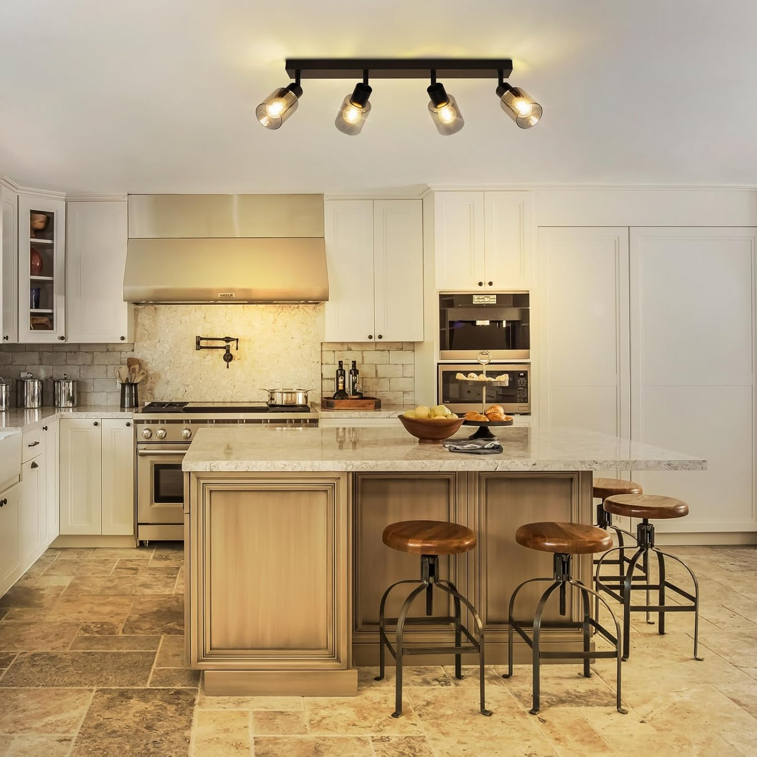 Kuchnia z wyspą, stołkami i oświetleniem sufitowym. Białe szafki, jasna wyspa i kamienna podłoga.