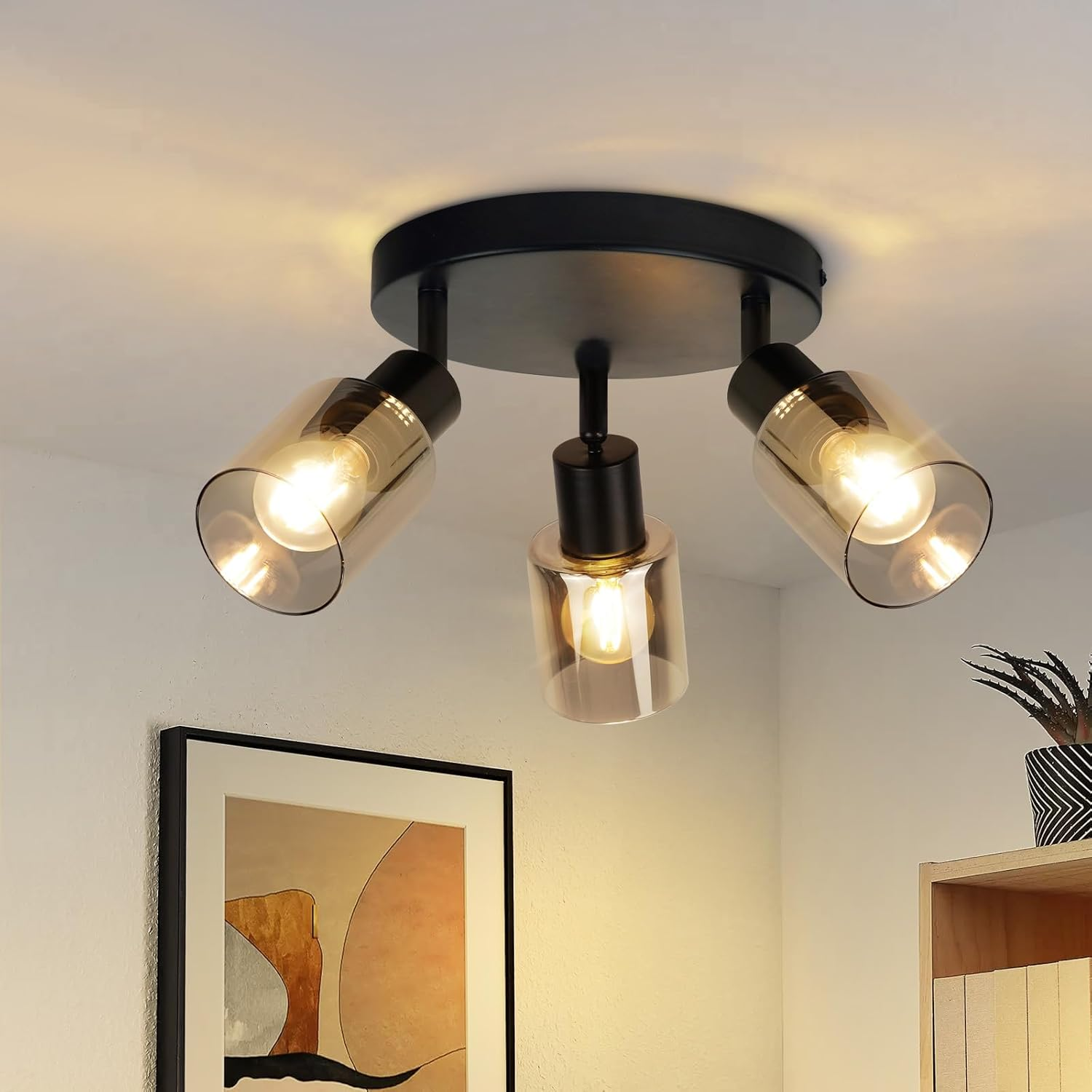 Lampa sufitowa z trzema abażurami. Obraz i roślina są w tle.
