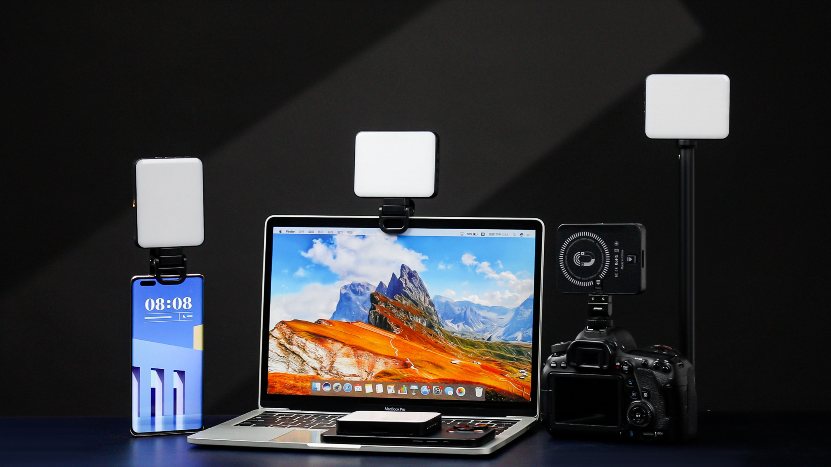 Laptop, Telefon, Kamera und LED-Leuchten auf dunkler Oberfläche. Telefon zeigt Uhrzeit. Lichter sind weiß und an.