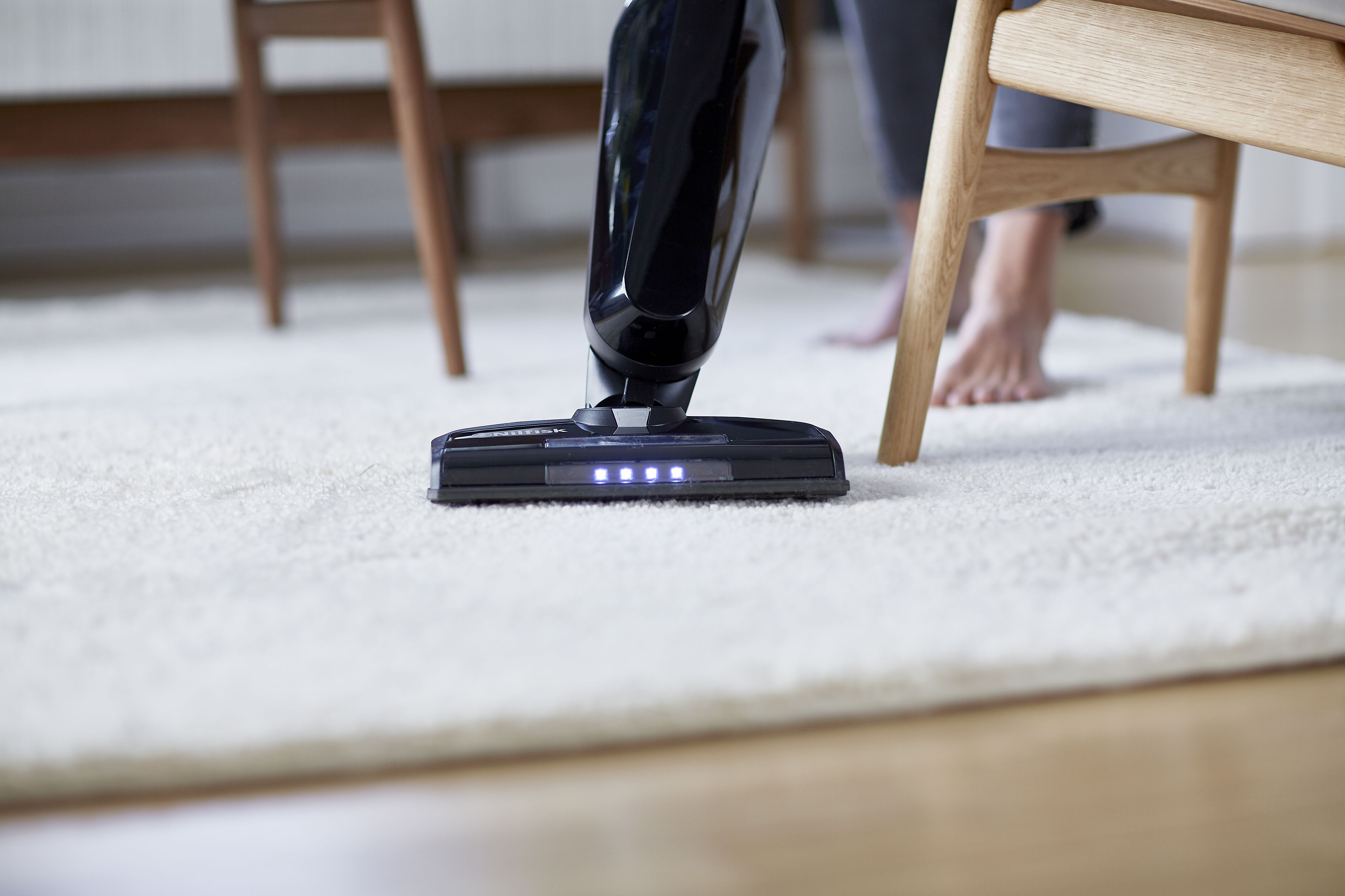 Aspirateur nettoyant un tapis blanc, avec les pieds d'une personne en arrière-plan.