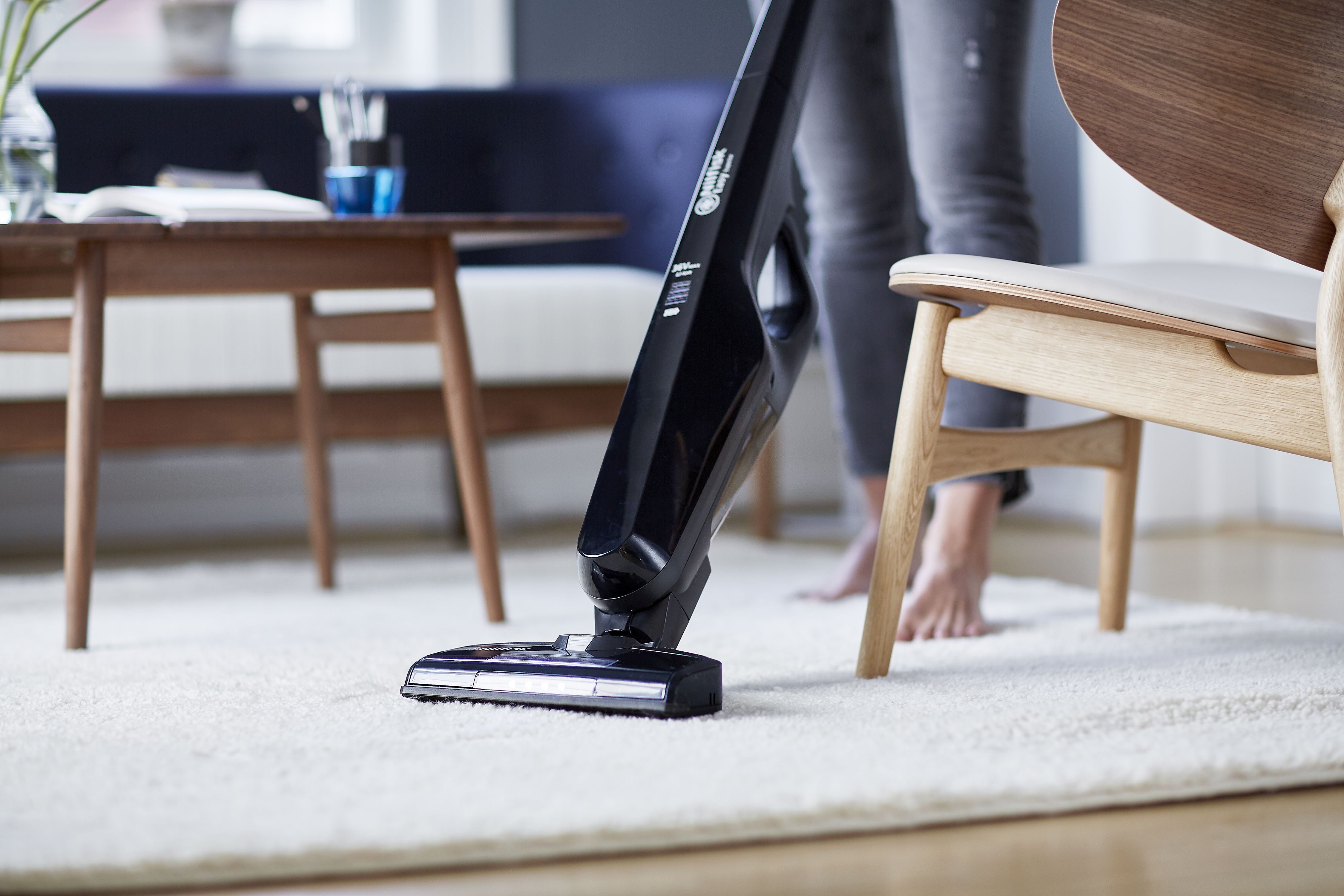 Une personne passant l'aspirateur sur un tapis blanc avec un aspirateur sans fil noir. Meubles en bois présents.