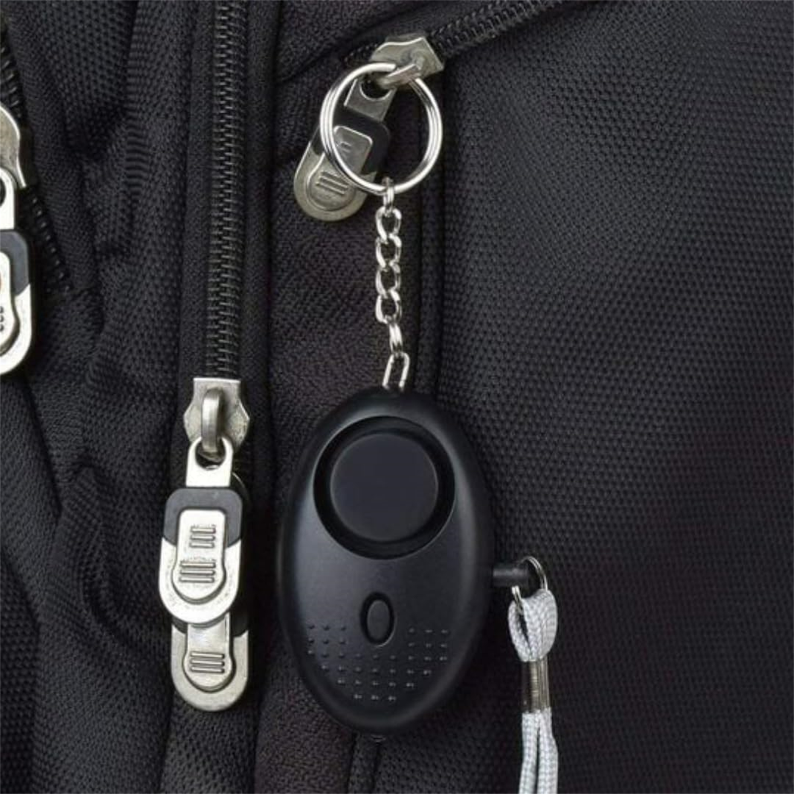Ein schwarzer persönlicher Alarm an einem Schlüsselanhänger, befestigt an einem schwarzen Rucksack. Er hat einen Knopf und eine kleine Kette.