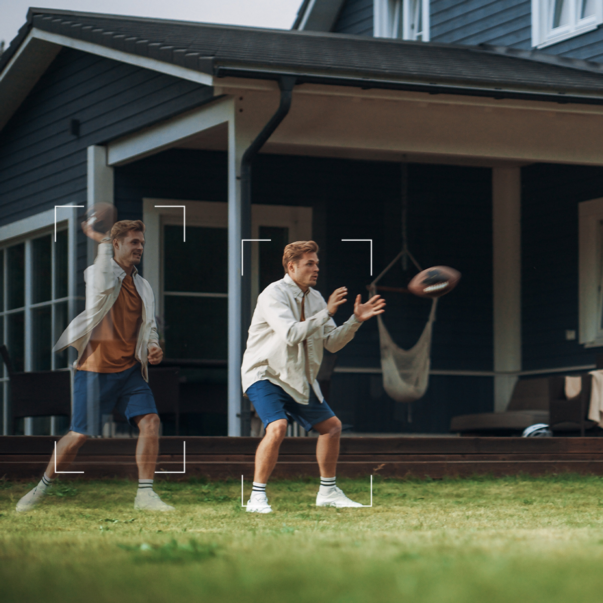 Dwóch mężczyzn grających w piłkę nożną na podwórku, jeden rzuca piłkę, dom w tle.