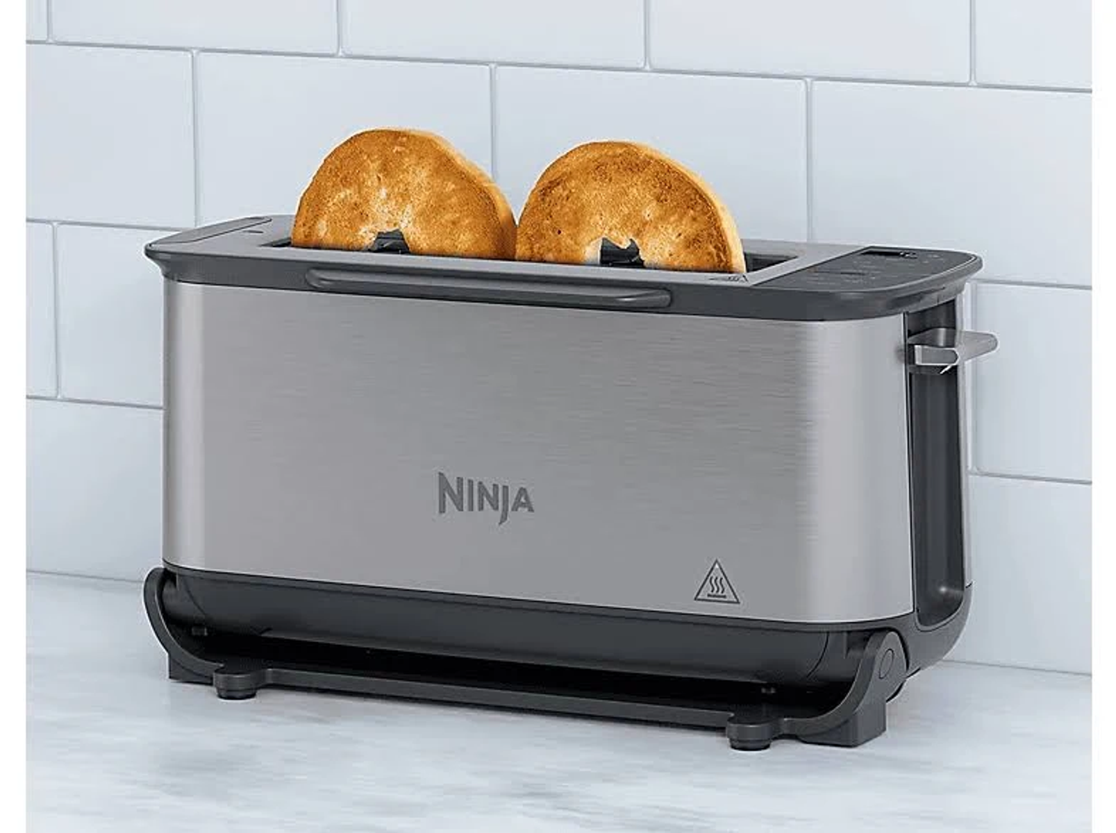 Toster Ninja ze stali nierdzewnej z dwoma tostowanymi bajglami w środku, białe kafelki w tle.