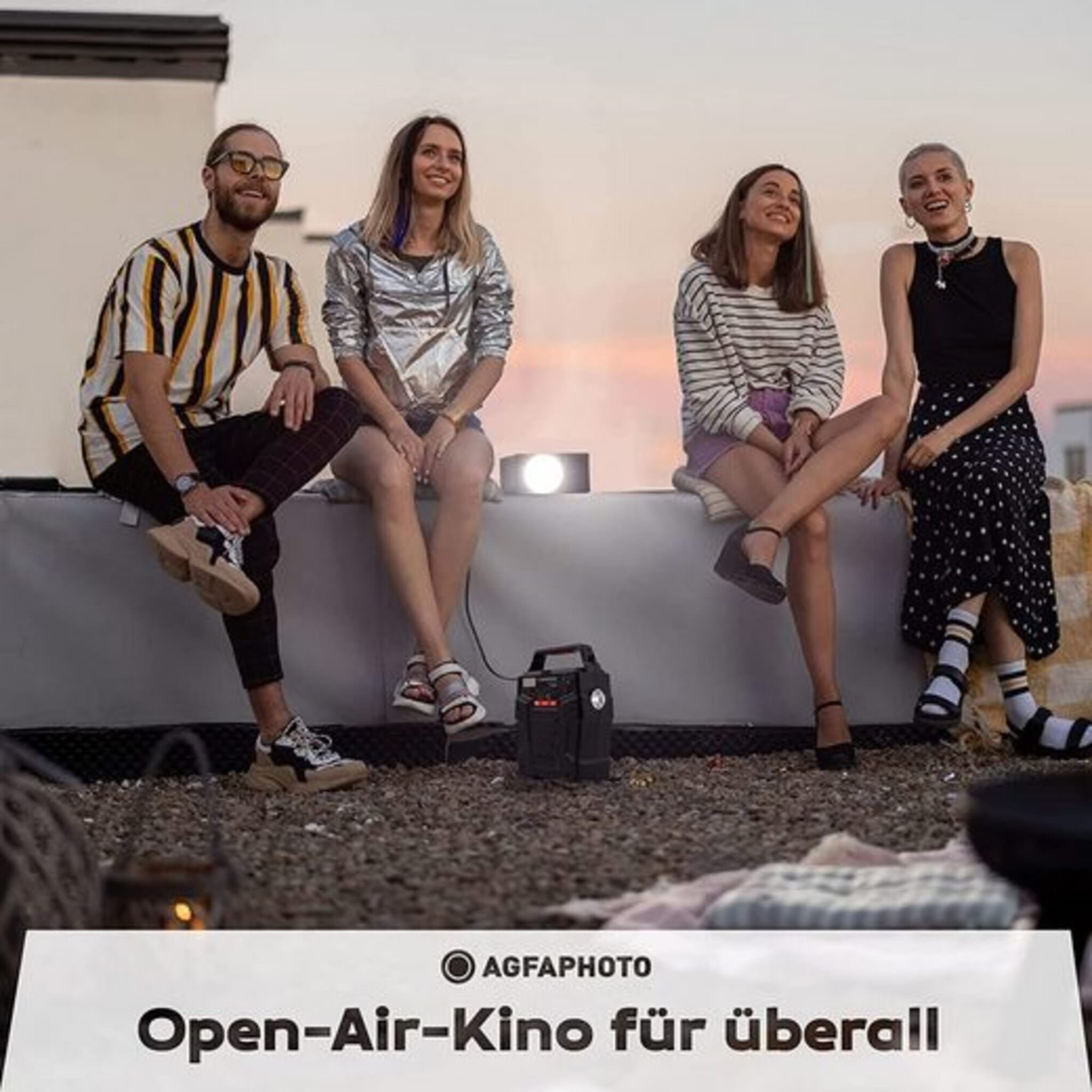 Vier Personen sitzen und schauen auf eine Projektorleinwand. Ein tragbarer Projektor steht auf dem Boden.