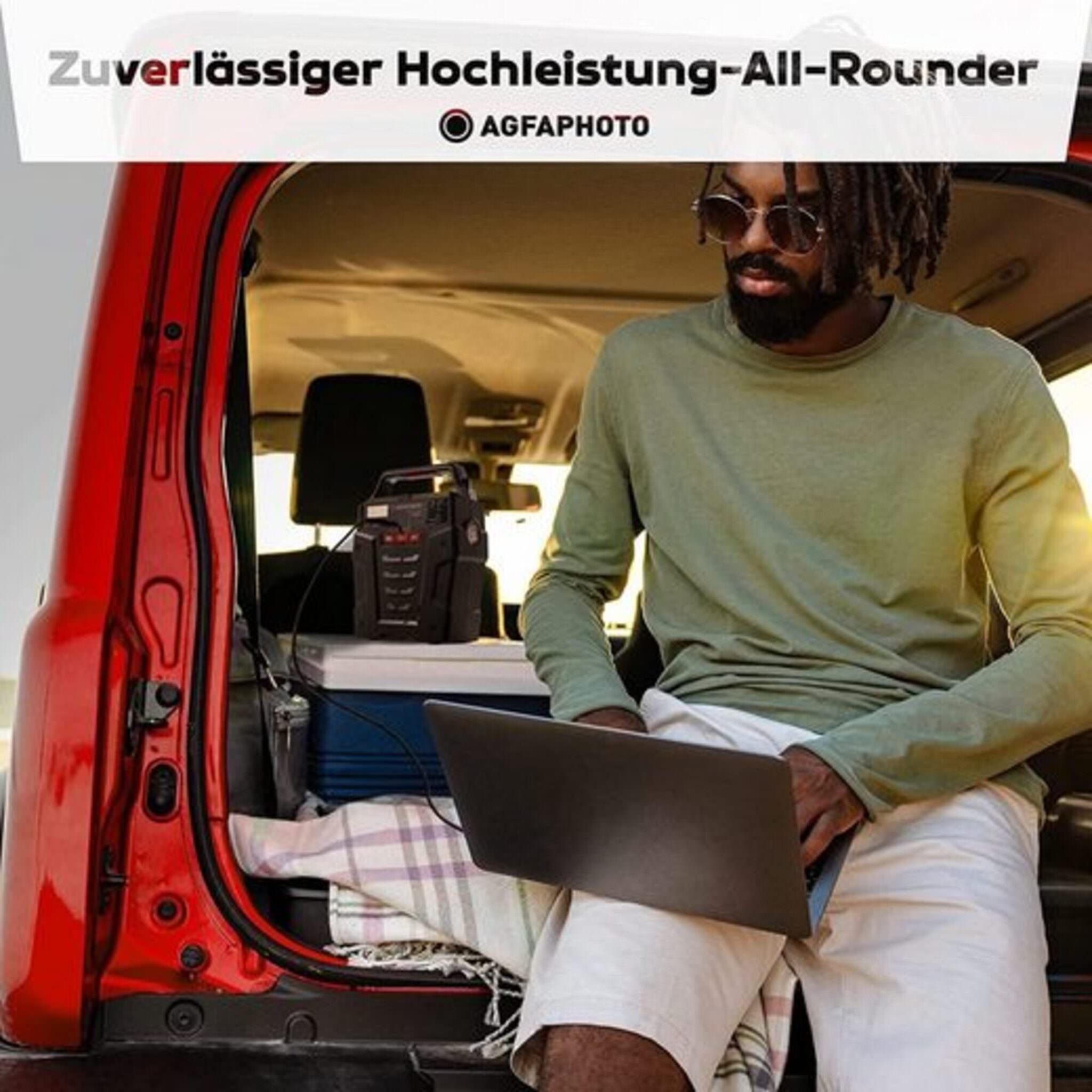 Ein Mann arbeitet mit einem Laptop im Heck eines roten Autos mit offener Hecktür, vor einem Strand.