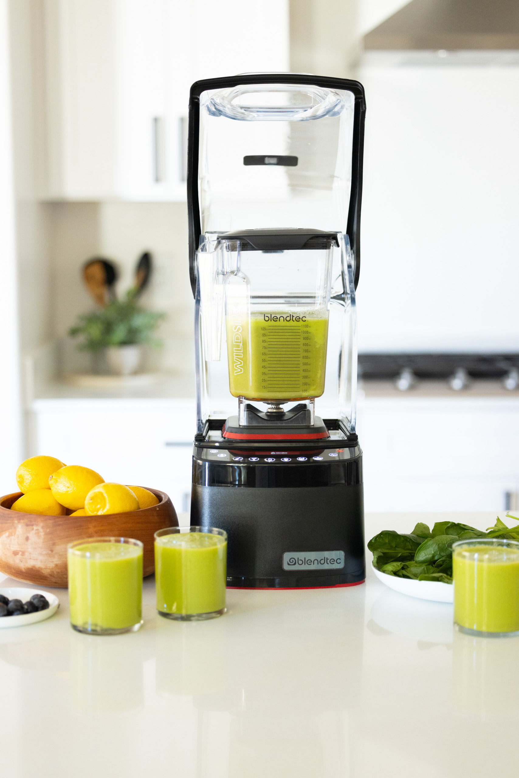 Frullatore con liquido verde, ciotola di limoni, bicchieri e verdure su bancone.