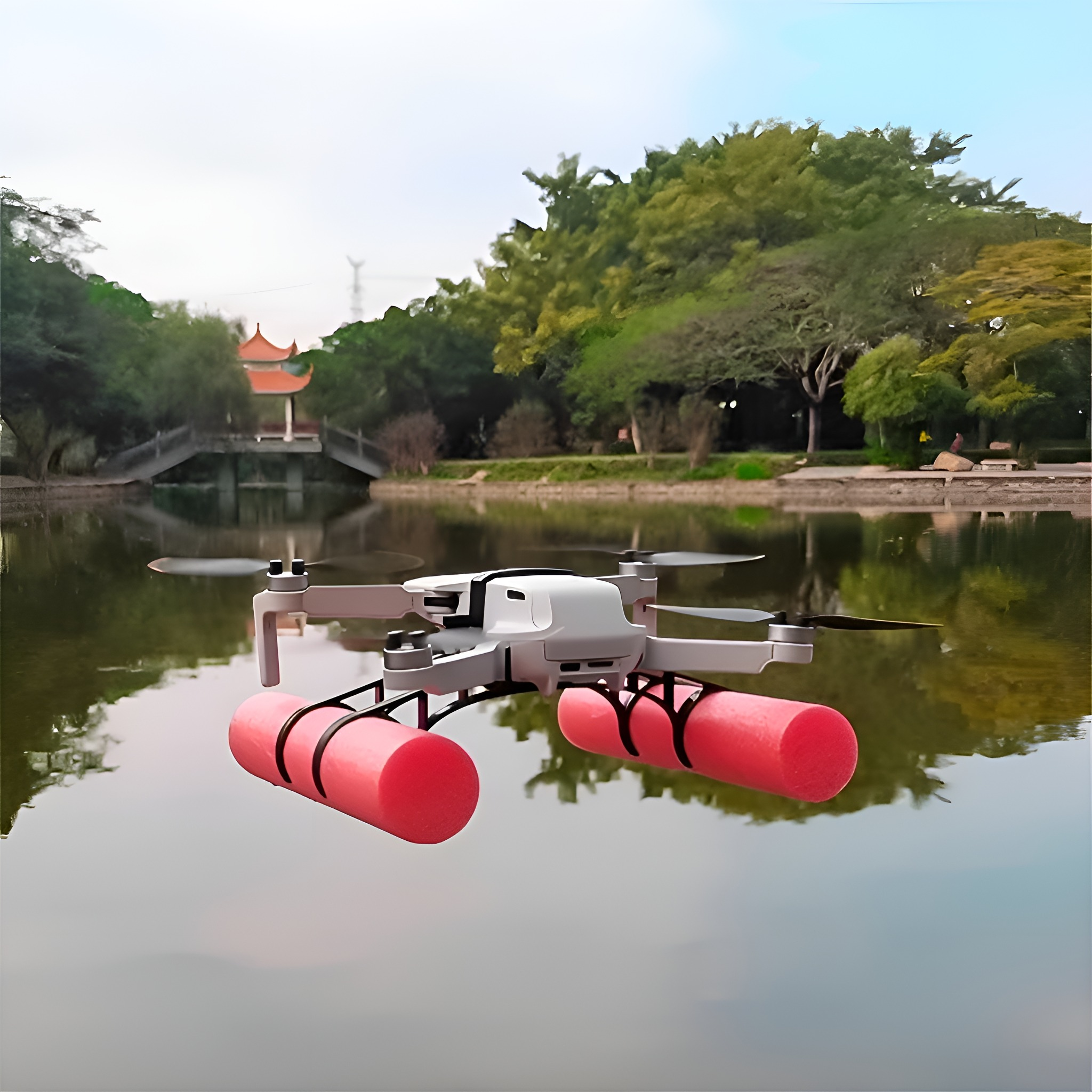 Eine Drohne schwebt über Wasser mit zwei rosa Schwimmern. Im Hintergrund befinden sich eine Brücke und Bäume.