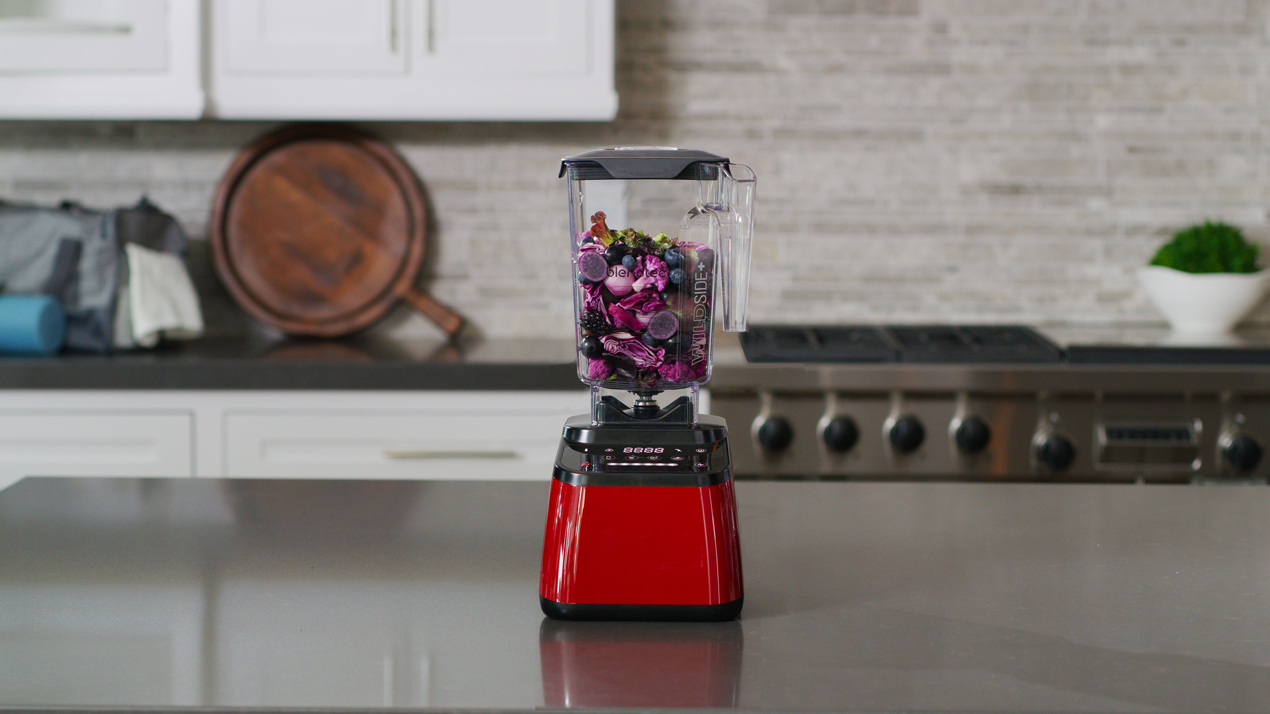 Frullatore rosso con ingredienti all'interno. È su un piano di lavoro. Lo sfondo mostra una cucina.