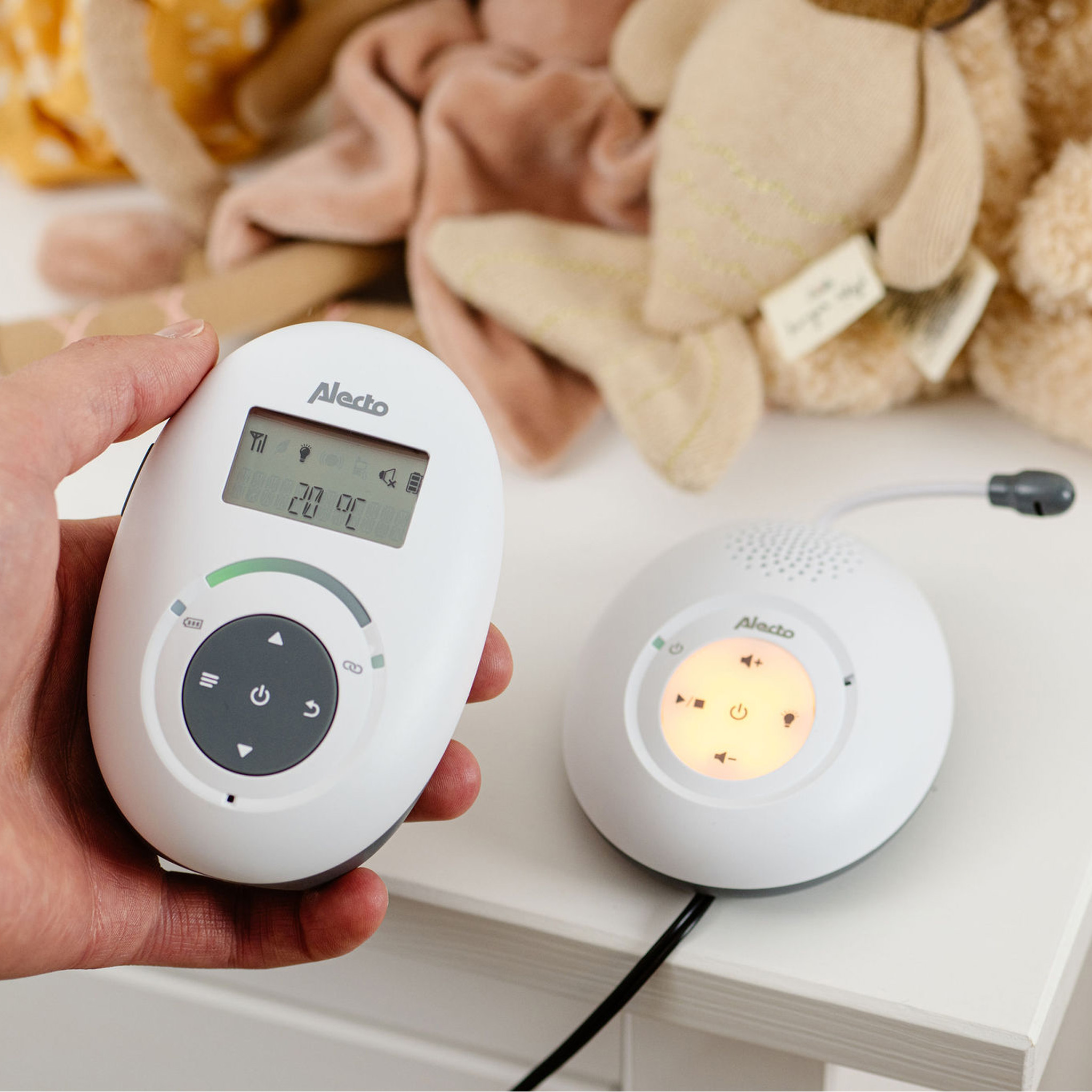 Ein Babyphone: ein weißer Monitor in der Hand und ein weißer Empfänger auf einem Tisch mit Stofftieren.