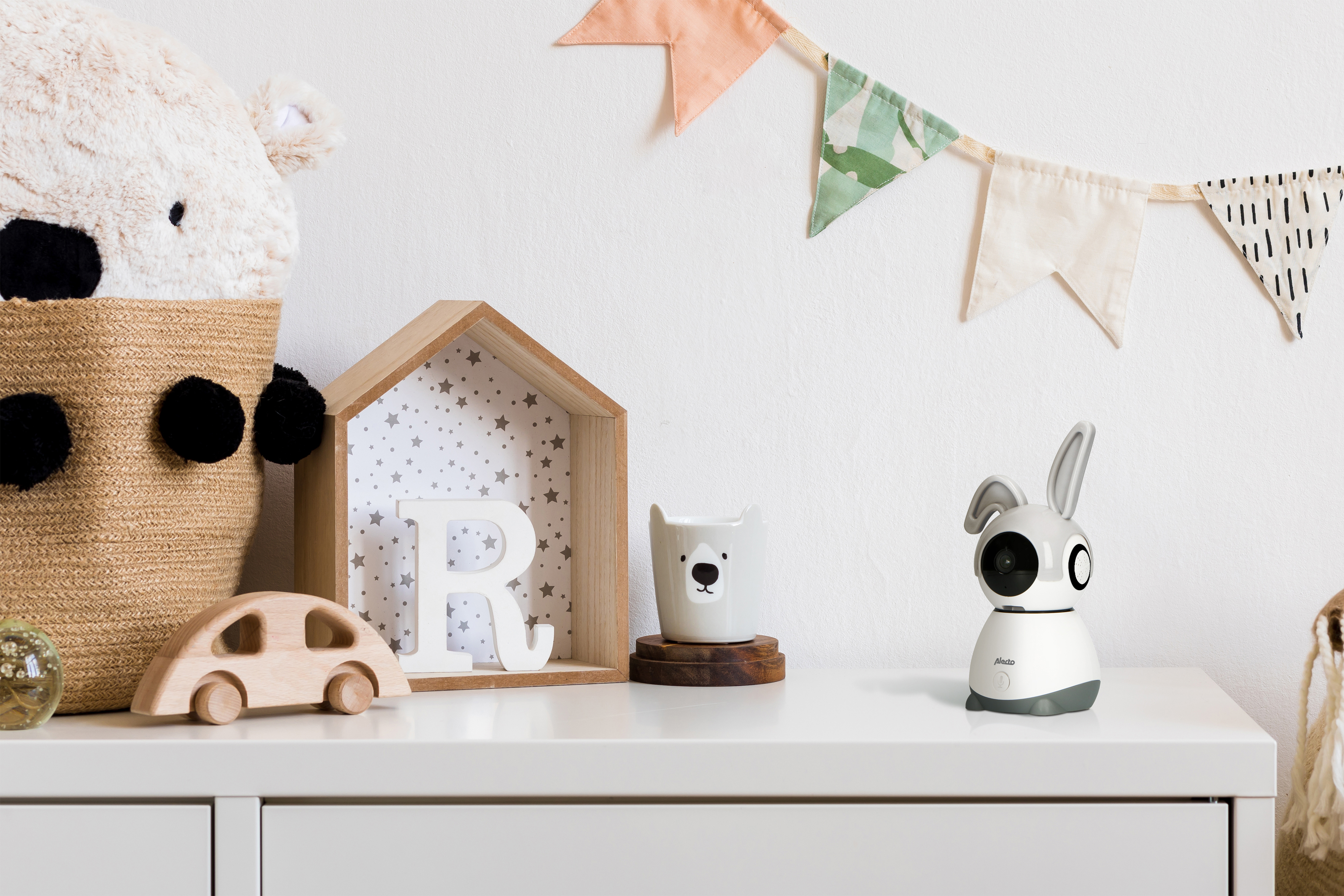 Babyphone auf weißer Oberfläche. Es ist weiß und grau, mit einer kameraförmigen Kamera. Spielzeug im Hintergrund.