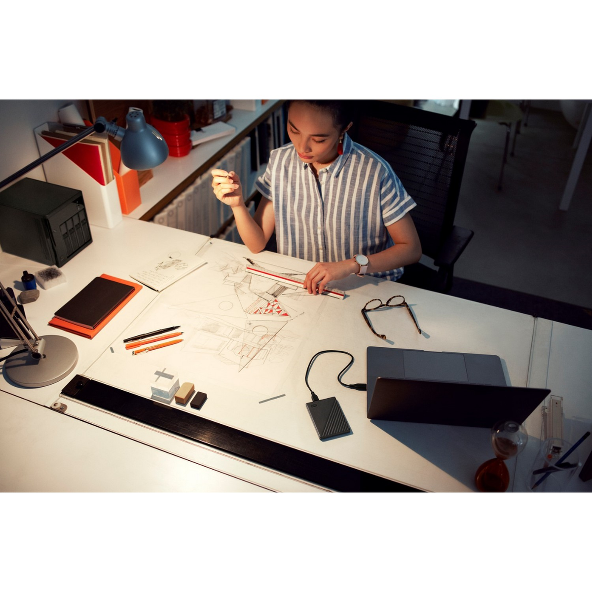 Kobieta pracuje przy biurku z planami, laptopem i różnymi narzędziami do projektowania.