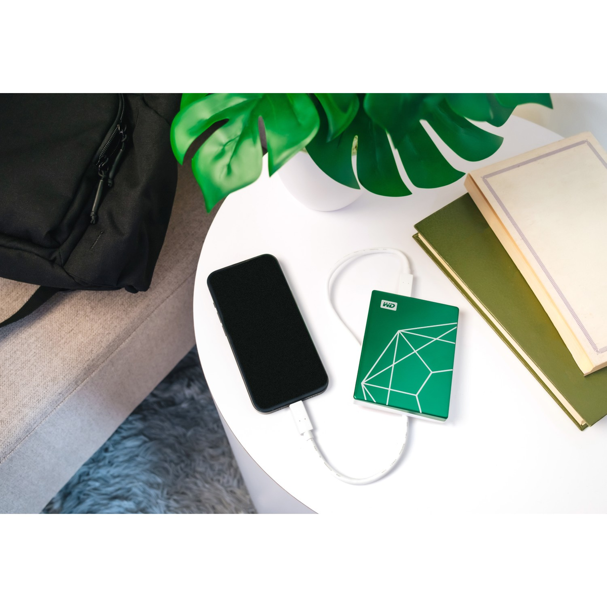 Telefon podłączony do zielonego przenośnego dysku twardego na stole. W pobliżu książki i roślina.