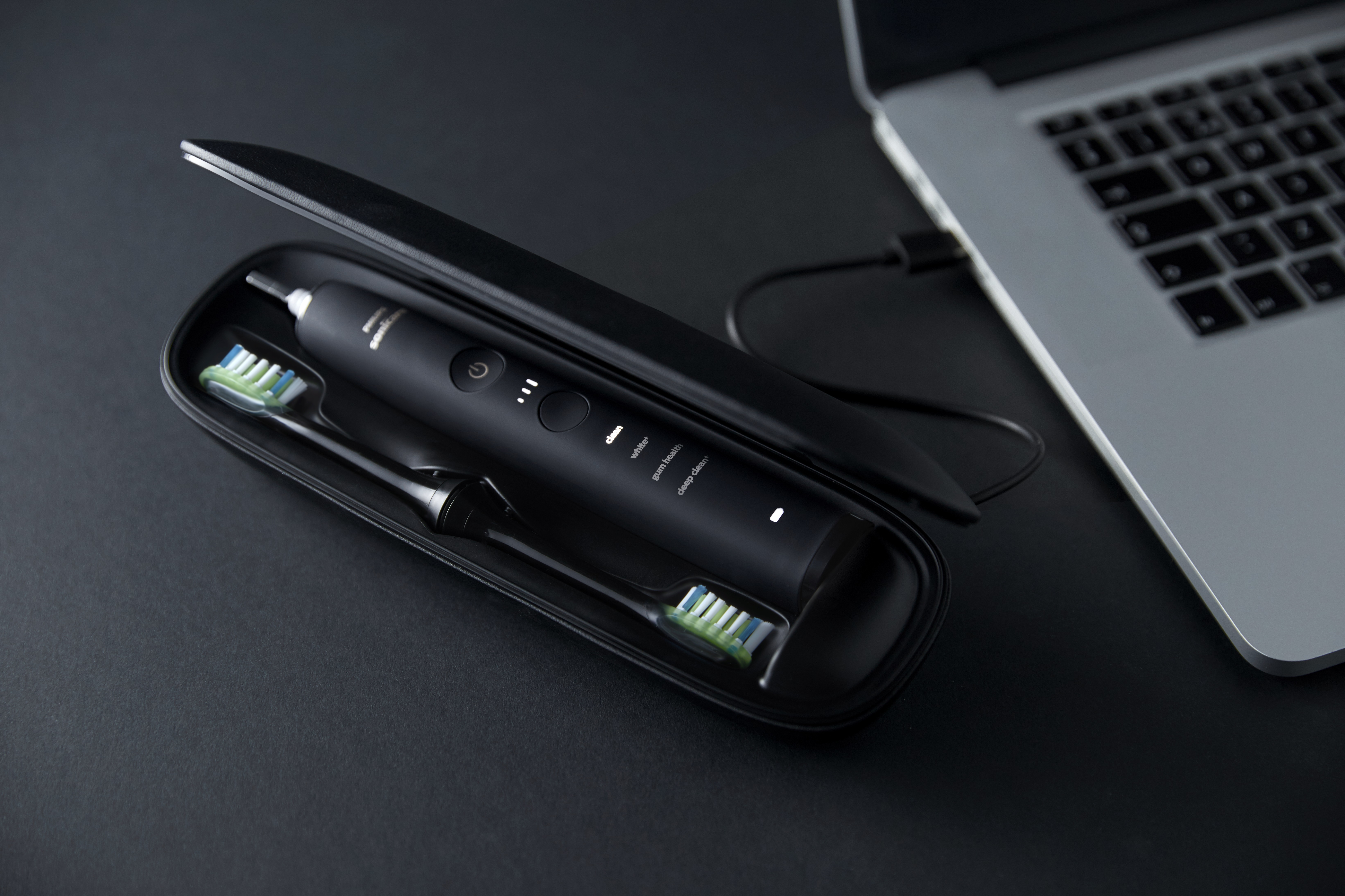 Black electric toothbrush kit in a case next to a laptop. There are two brush heads and a USB cable.