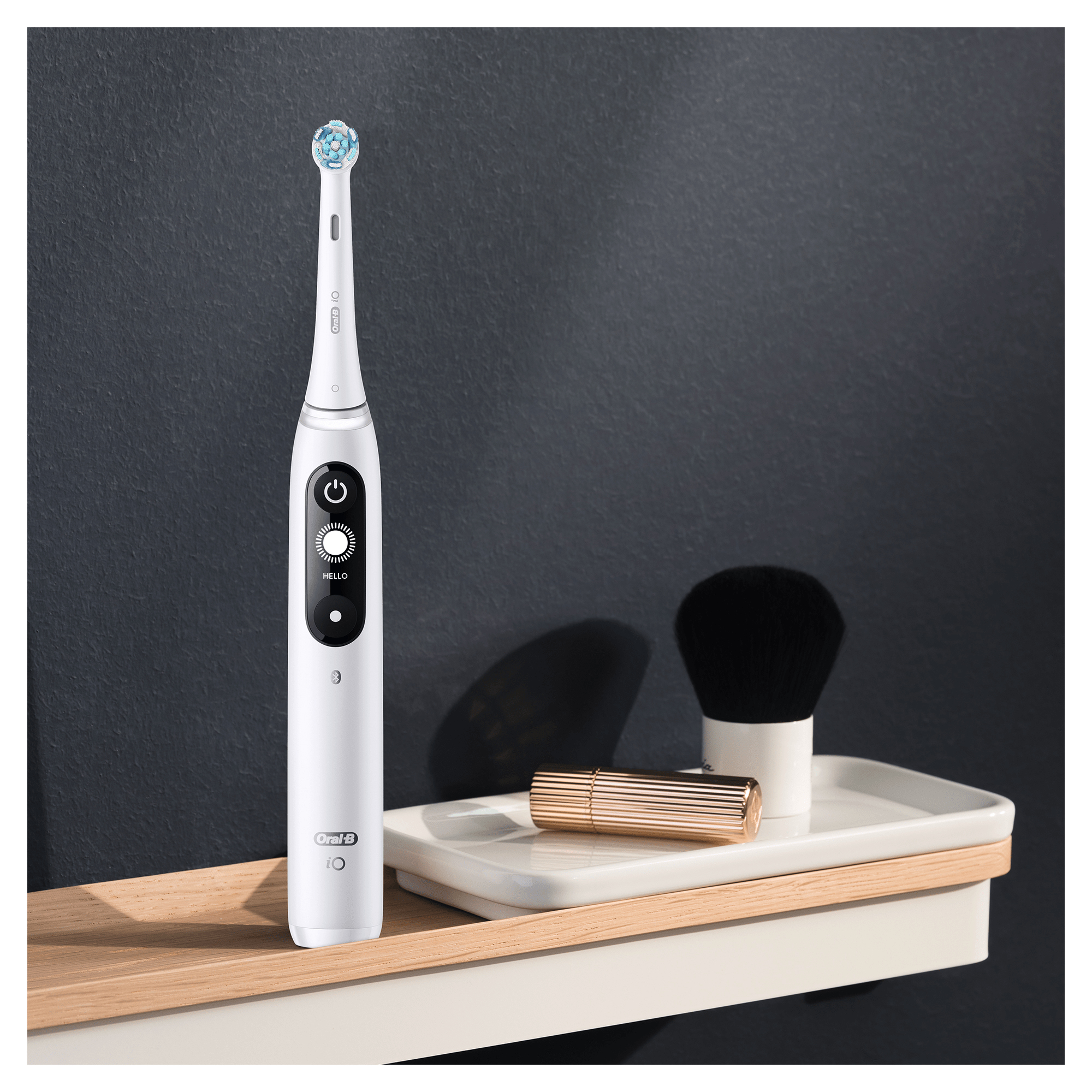 Spazzolino elettrico Oral-B bianco, su una mensola di legno. Un piccolo vassoio con rossetto e pennello.