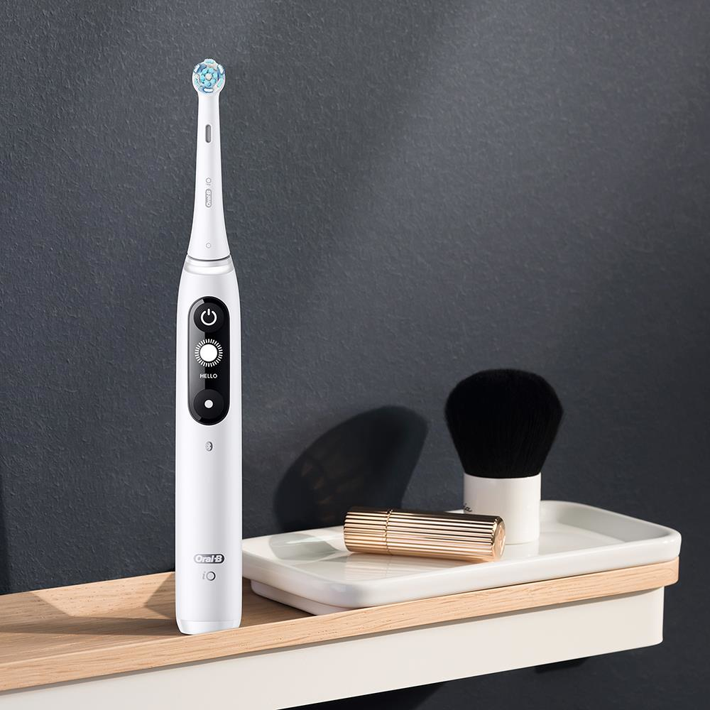 Un cepillo de dientes blanco Oral-B IO serie 6s en un estante con un cepillo y un lápiz labial. Fondo gris oscuro.