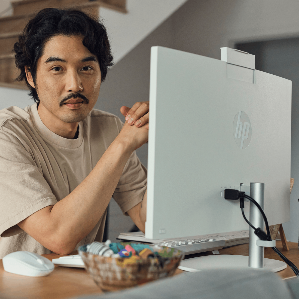 Hombre sentado en un escritorio, trabajando en una computadora blanca. Tiene un monitor, teclado, ratón y un cuenco con objetos.
