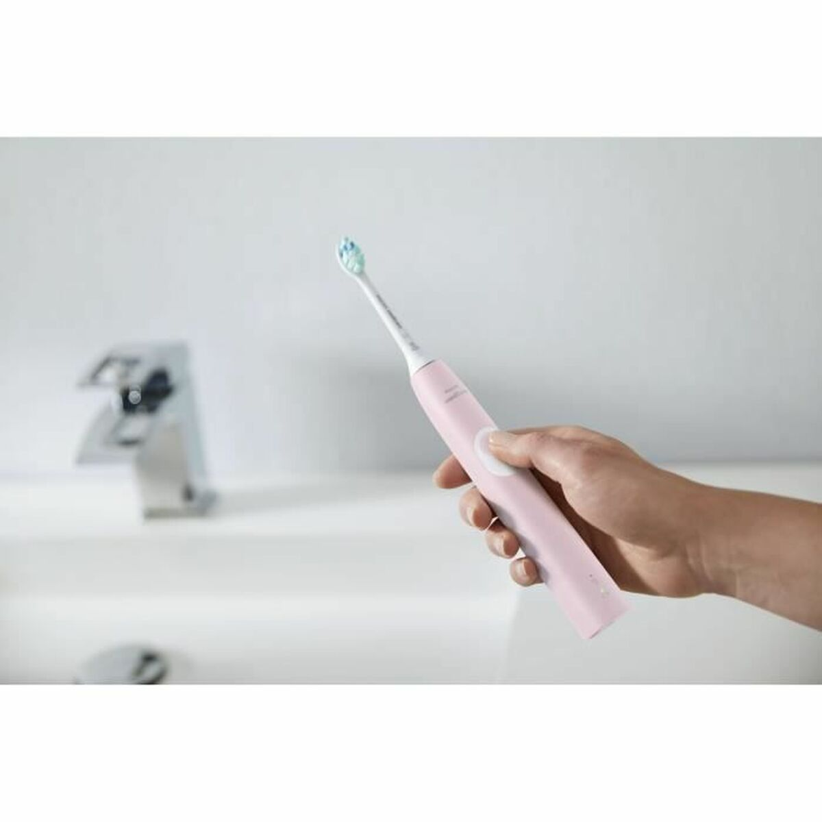 Persona che tiene uno spazzolino rosa vicino a un lavandino. Lo sfondo è un muro bianco e un rubinetto.