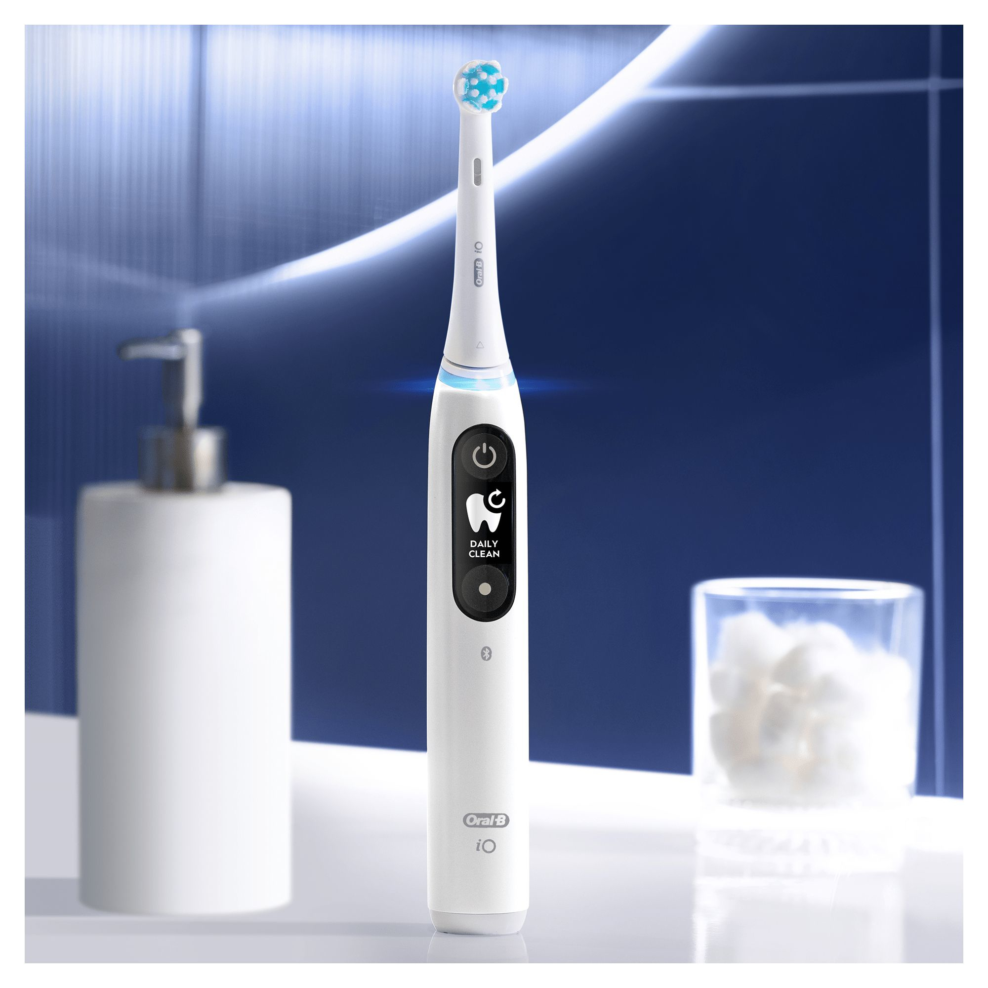 White electric toothbrush with black display. A white bottle and a glass of cotton balls are in the background.