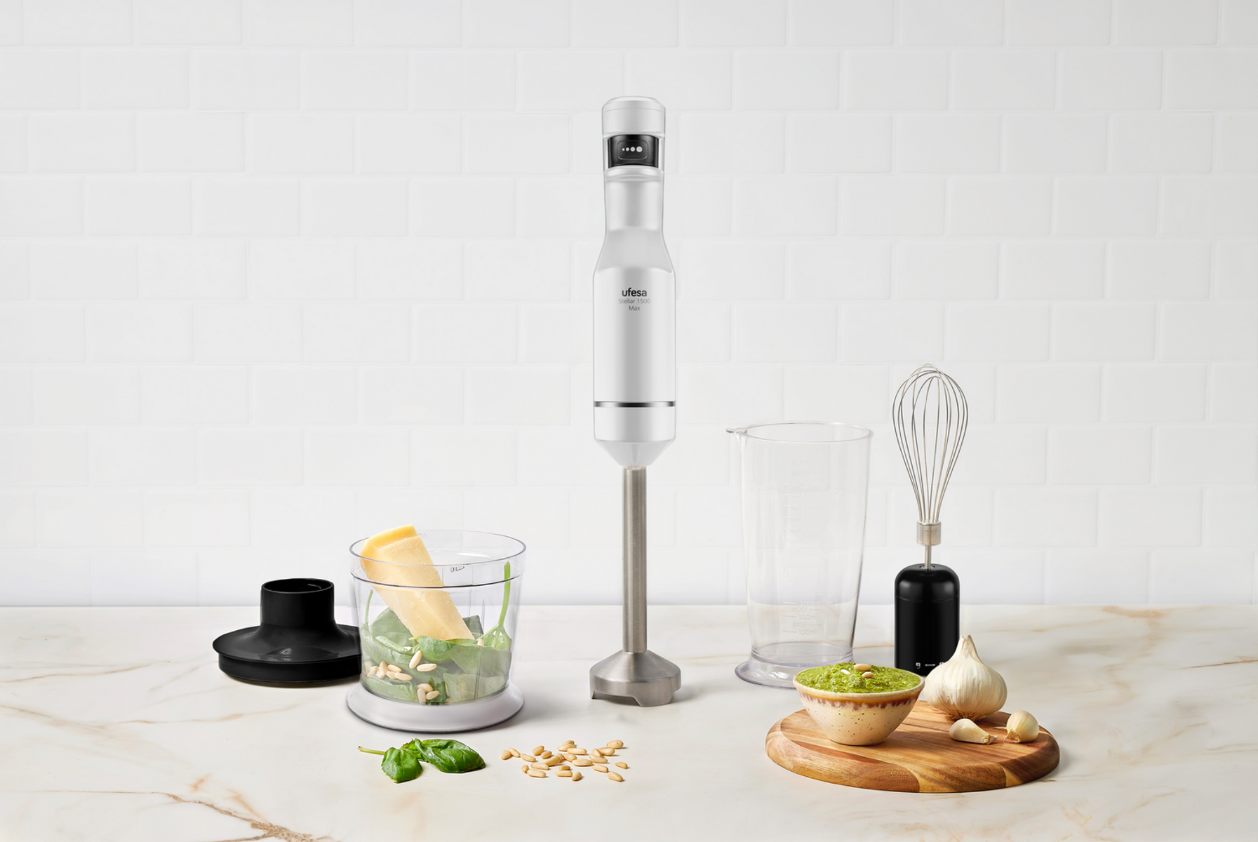 Un frullatore a immersione bianco con accessori. Include una ciotola con basilico, una frusta e uno spicchio d'aglio.