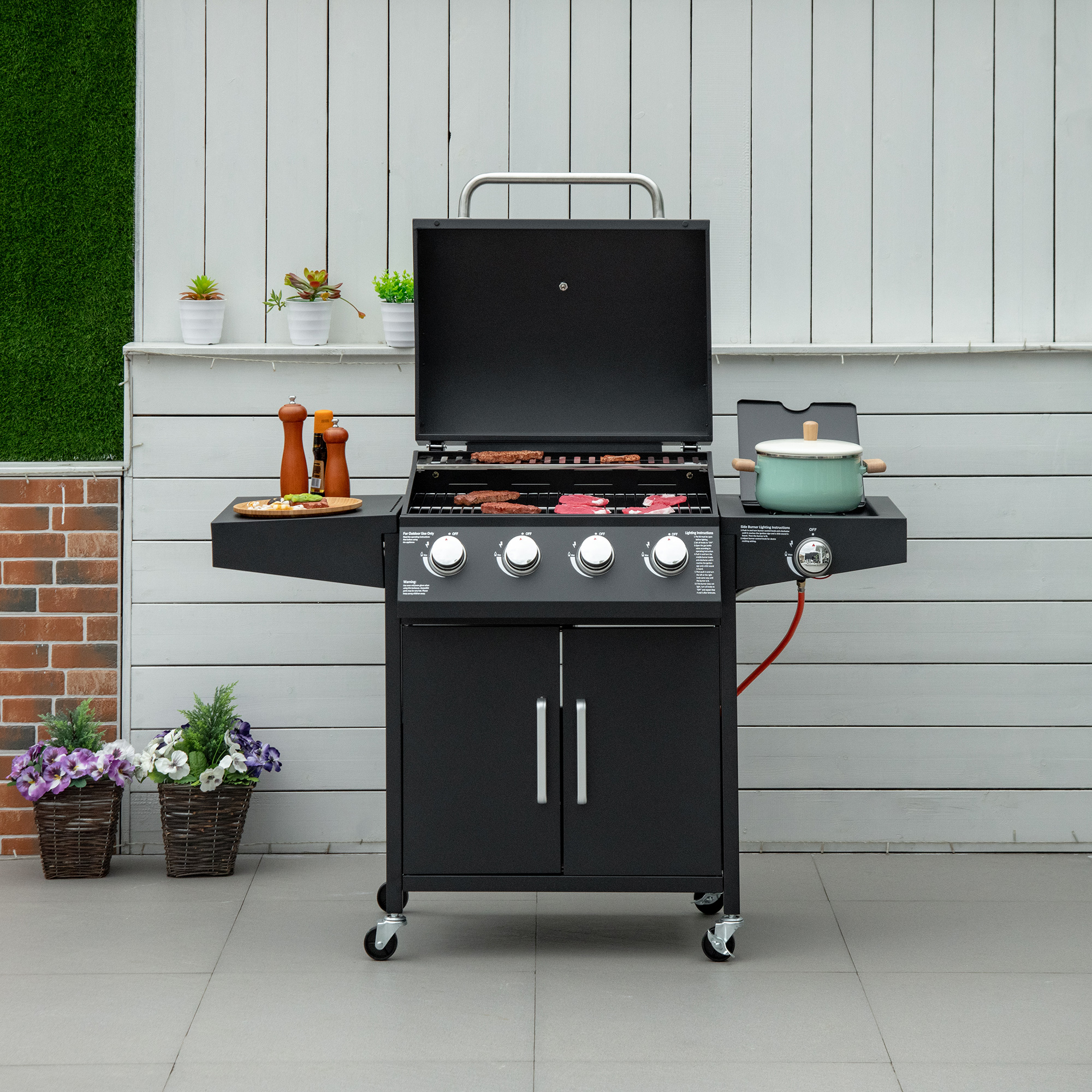 Schwarzer Außengrill mit offenem Deckel, Essen und Ablagen. Vor weißem Zaun.