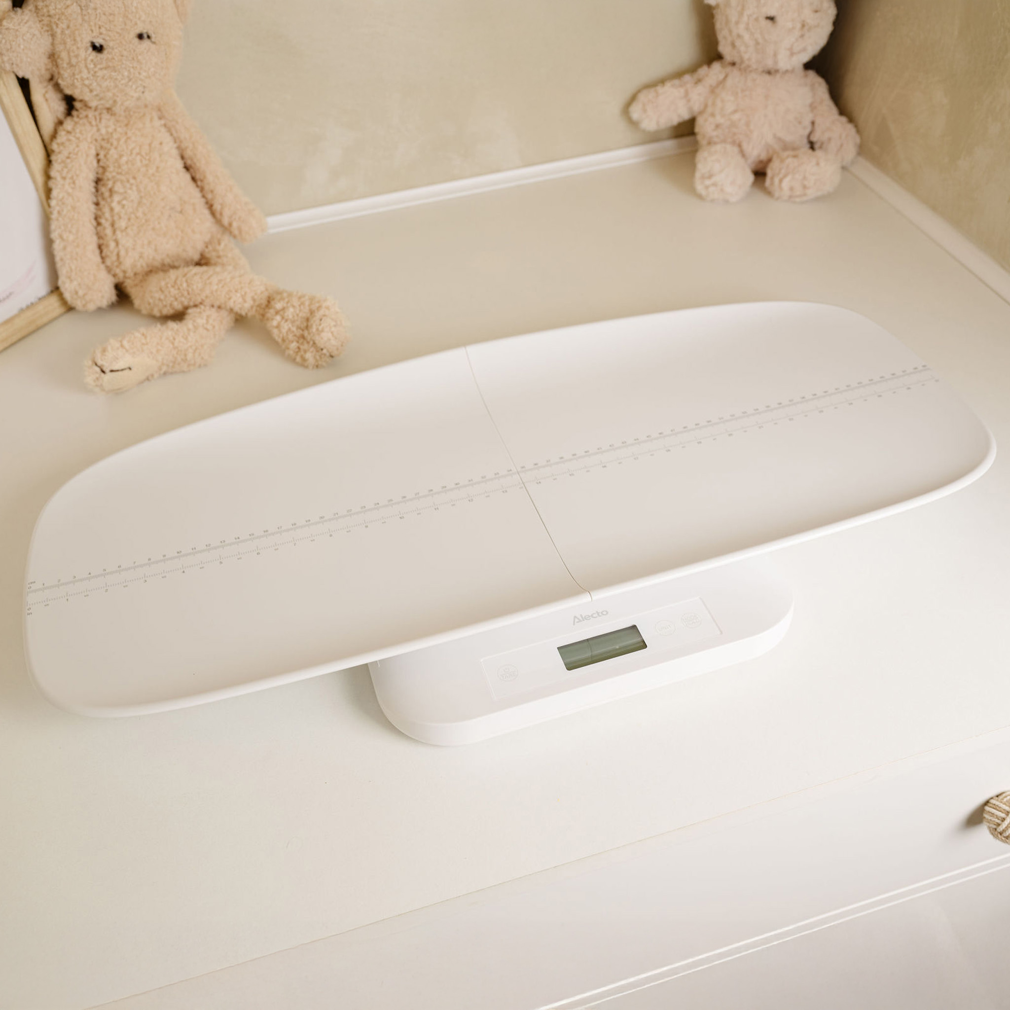 Weiße Babywaage auf weißer Oberfläche. Zwei Teddybären sitzen im Hintergrund.