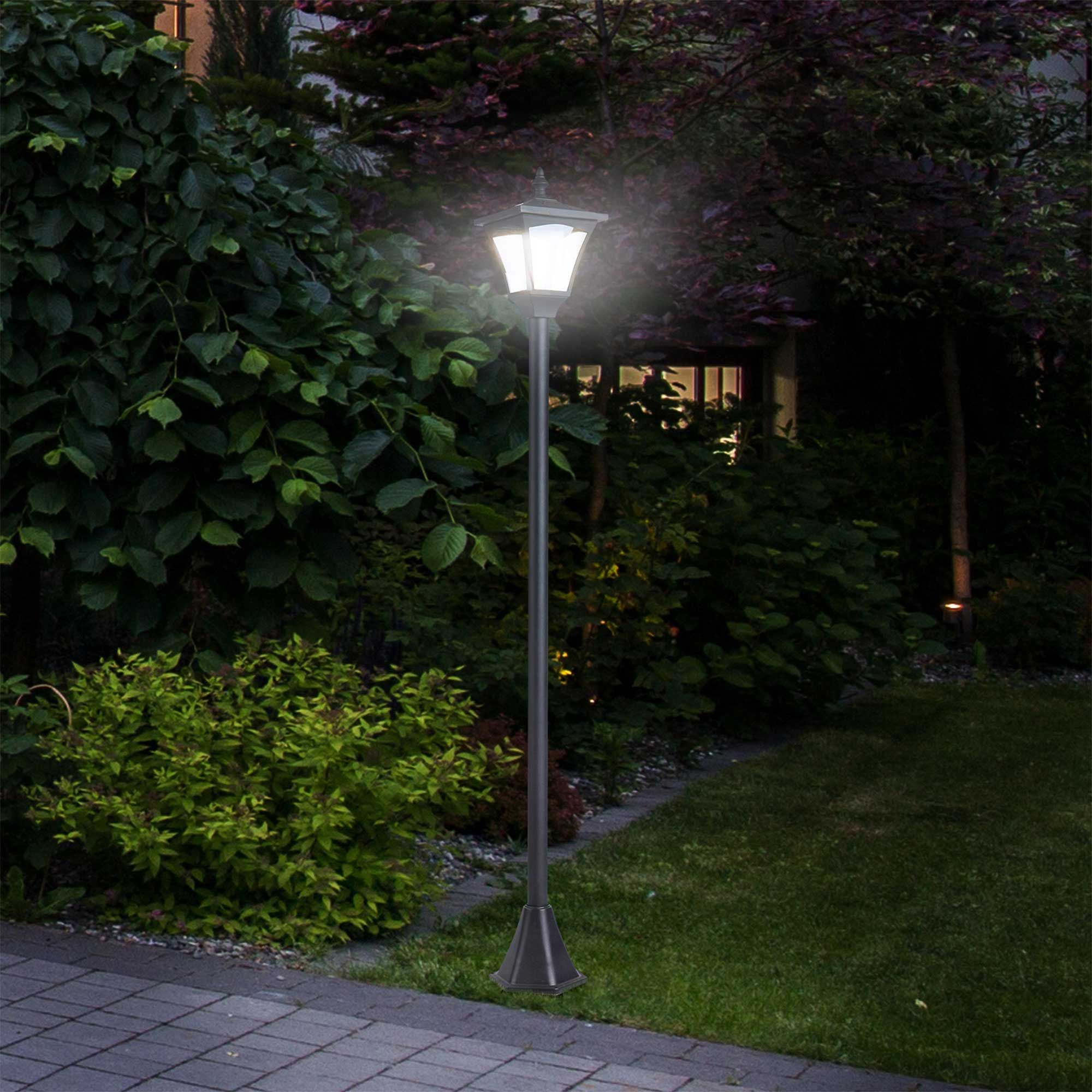 Szene im Freien mit einer beleuchteten Außenlampe in einem Garten. Büsche und Gras sind im Hintergrund.