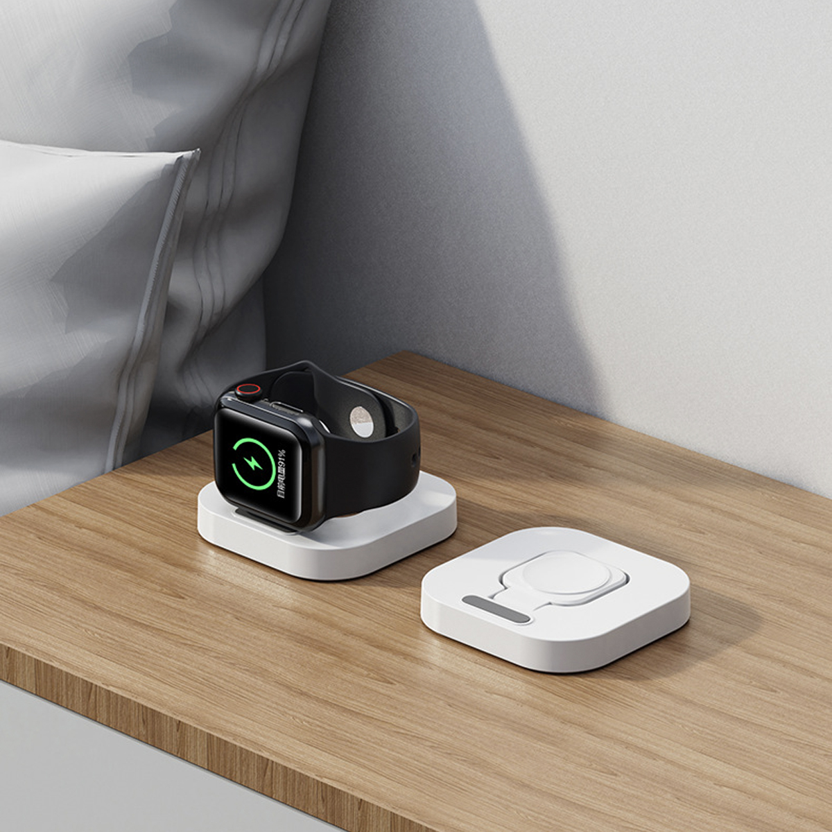Eine schwarze Smartwatch auf einem weißen Ladepad. Ein zweites Pad daneben. Beide auf einer Holzoberfläche, mit einem Kissen im Hintergrund.