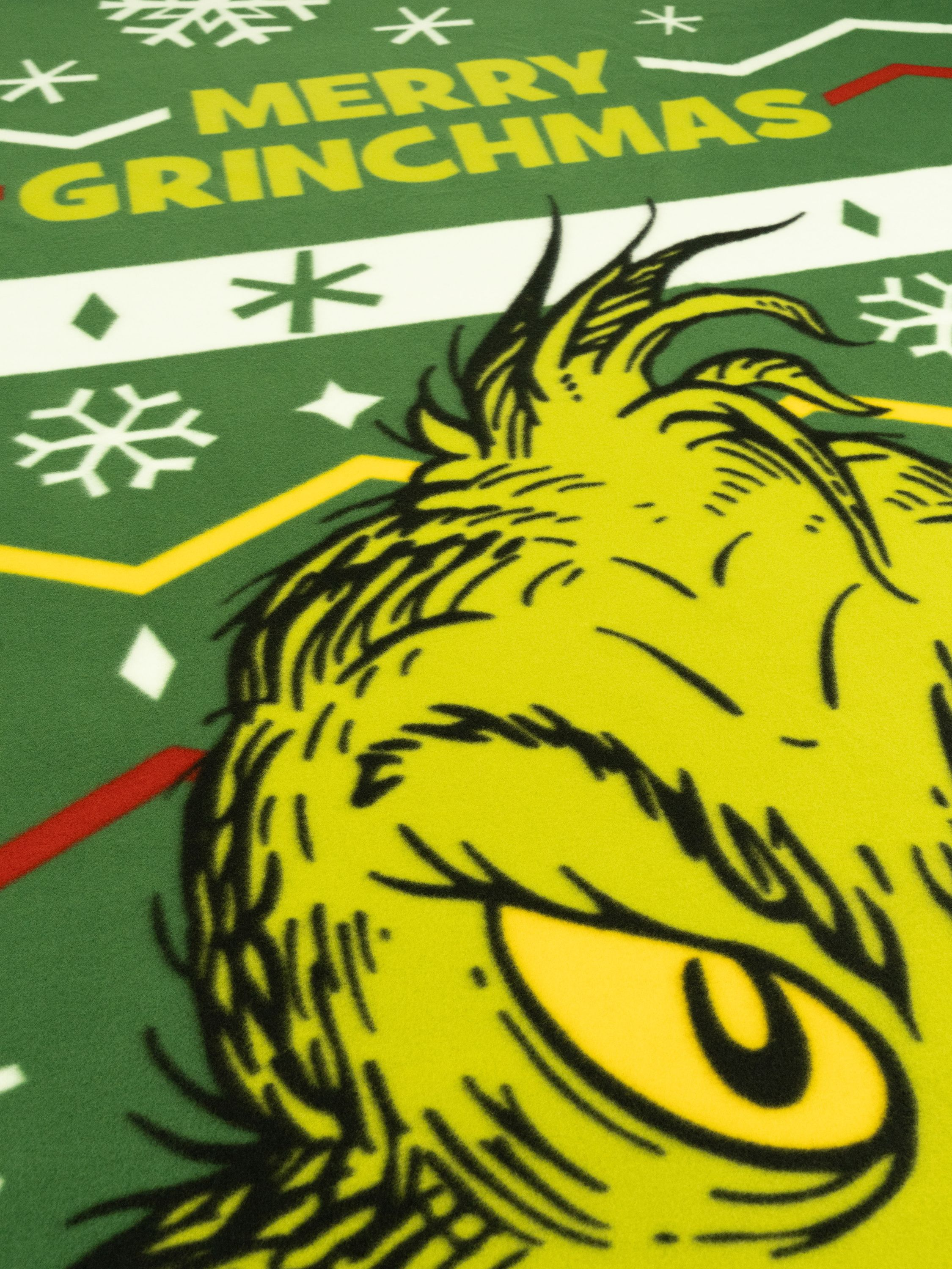 Nahaufnahme von Grinchs Gesicht auf grünem Hintergrund, mit "MERRY GRINCHMAS" Text und Schneeflocken.