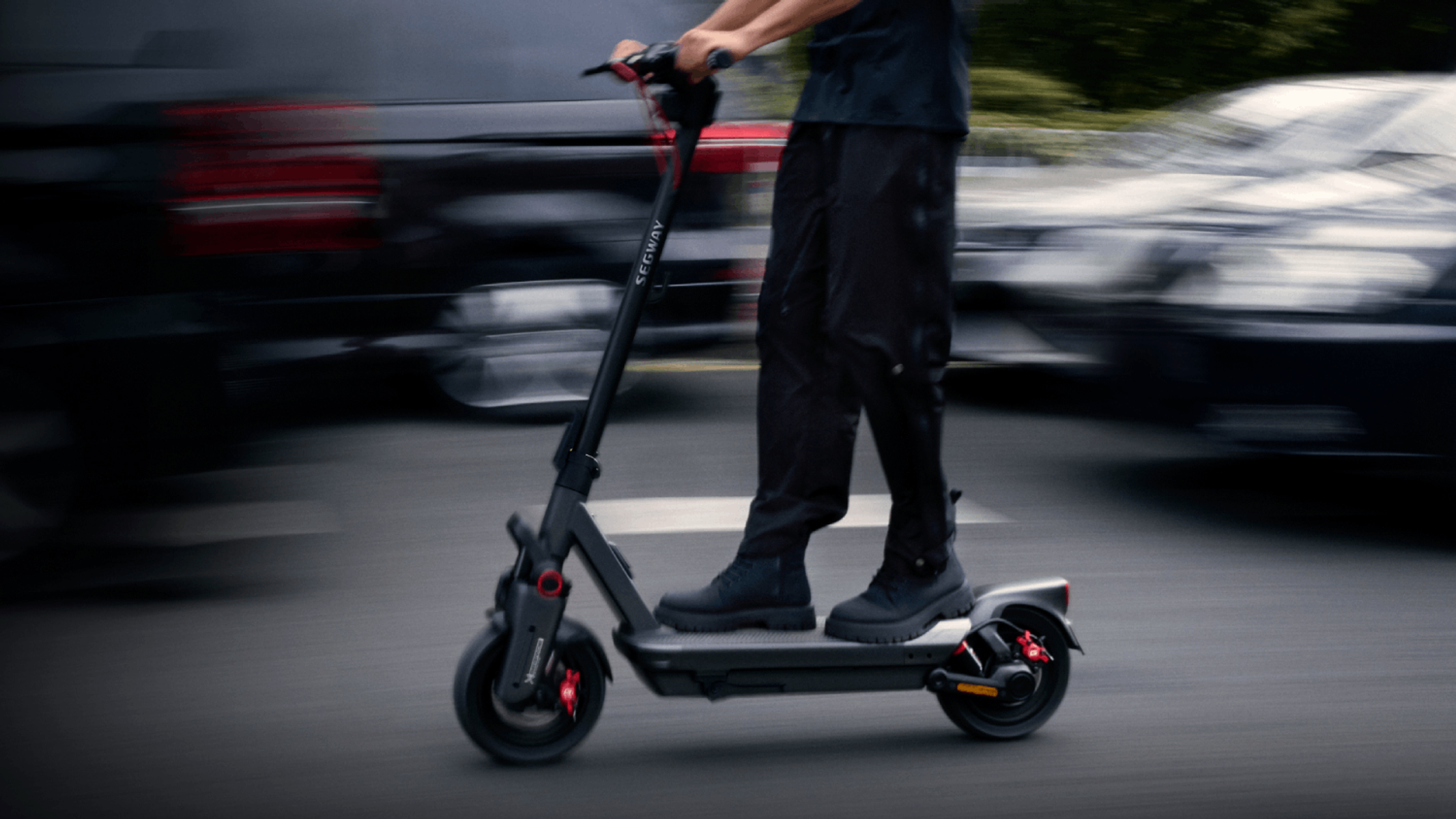 Persona che guida un monopattino elettrico nero e grigio su una strada. Sfondo sfocato con auto.