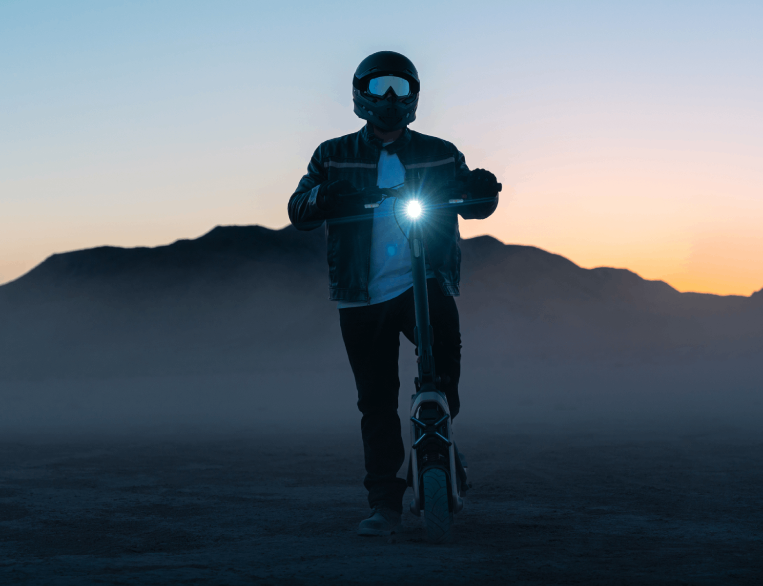 Person mit Helm und Roller in einer Wüstenlandschaft in der Abenddämmerung. Scheinwerfer ist an.