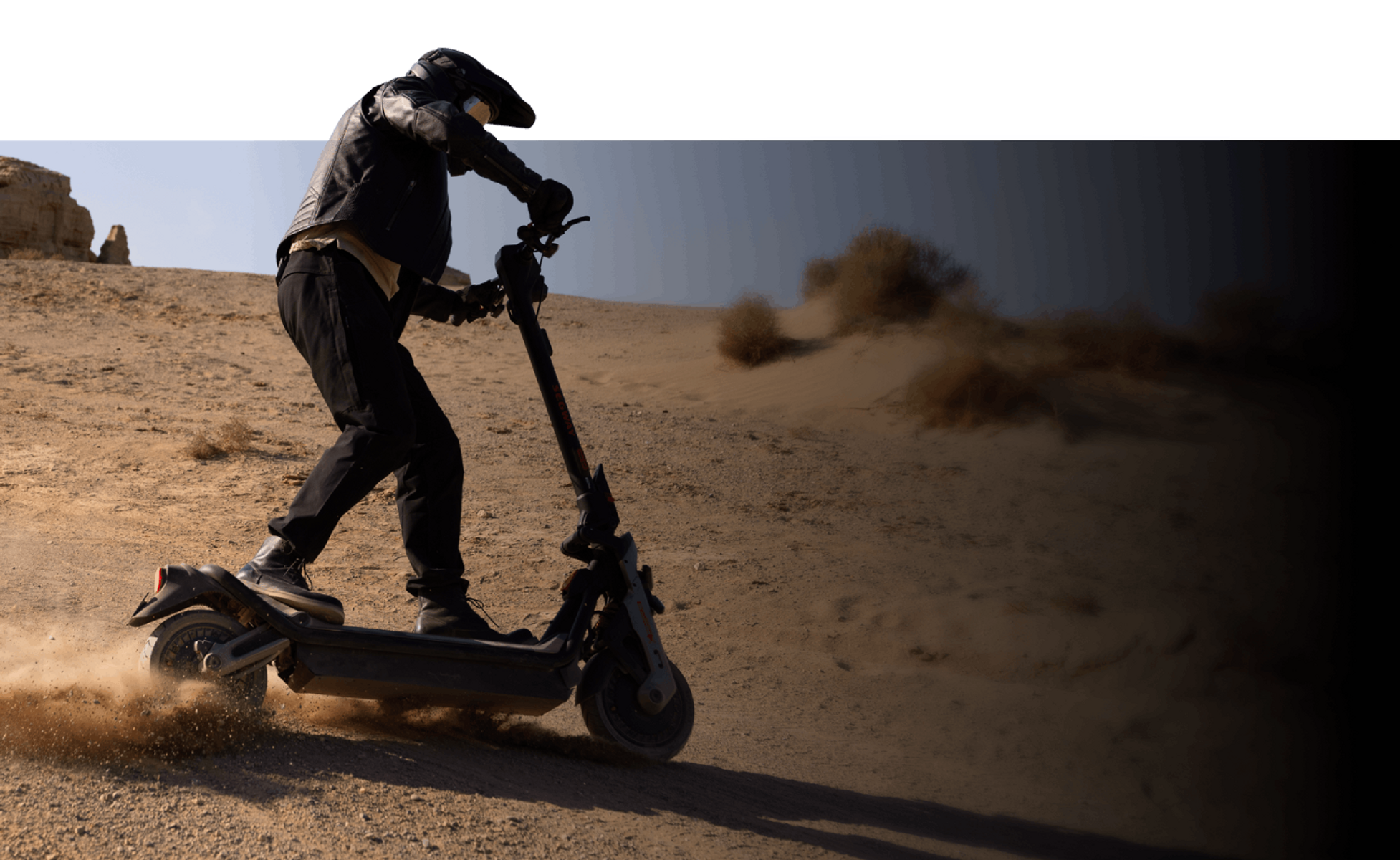 Osoba jedzie na hulajnodze elektrycznej po wydmie. Tło to pustynny krajobraz z jasnym niebem.