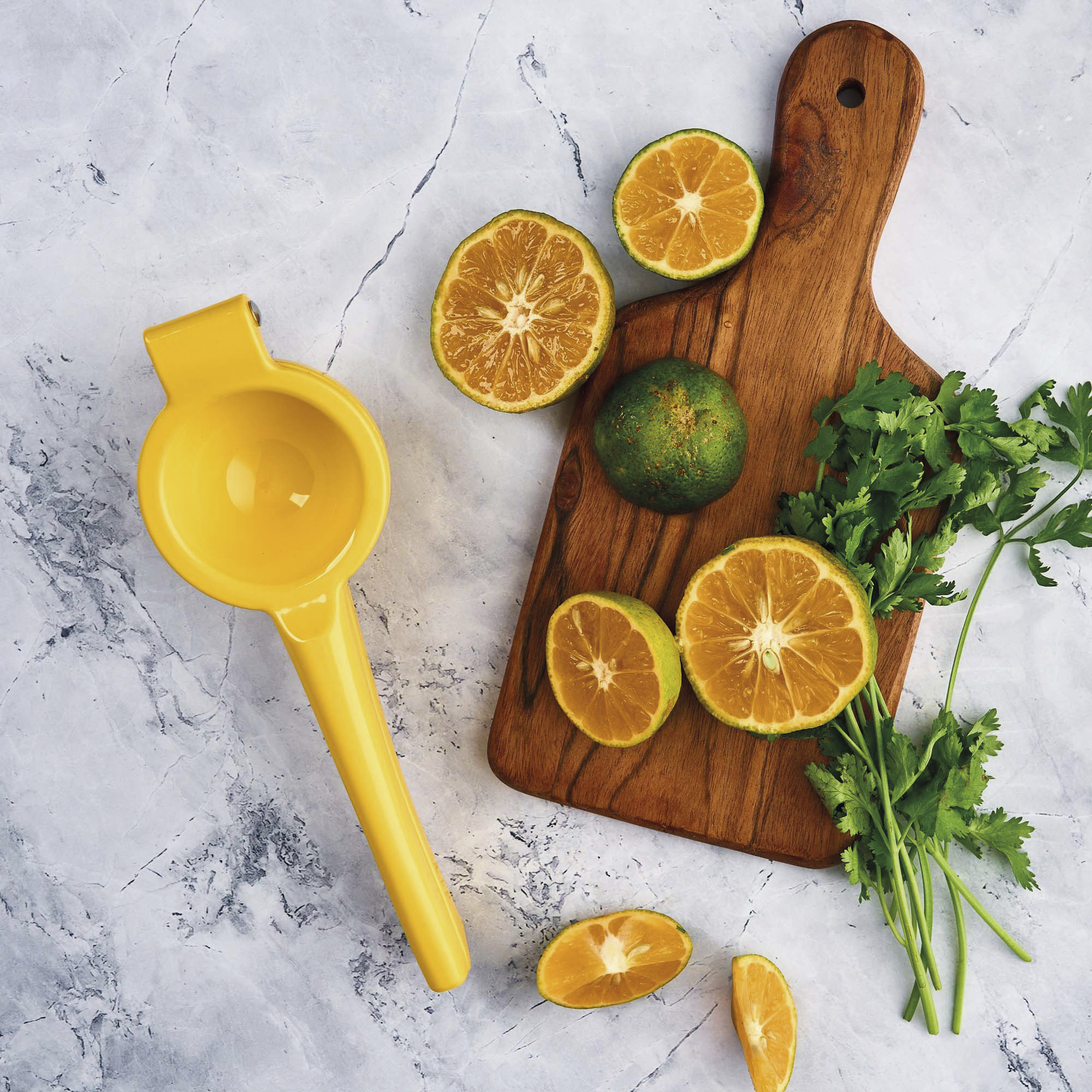 Exprimidor de cítricos amarillo con naranjas, cilantro y naranjas cortadas sobre una superficie de mármol.