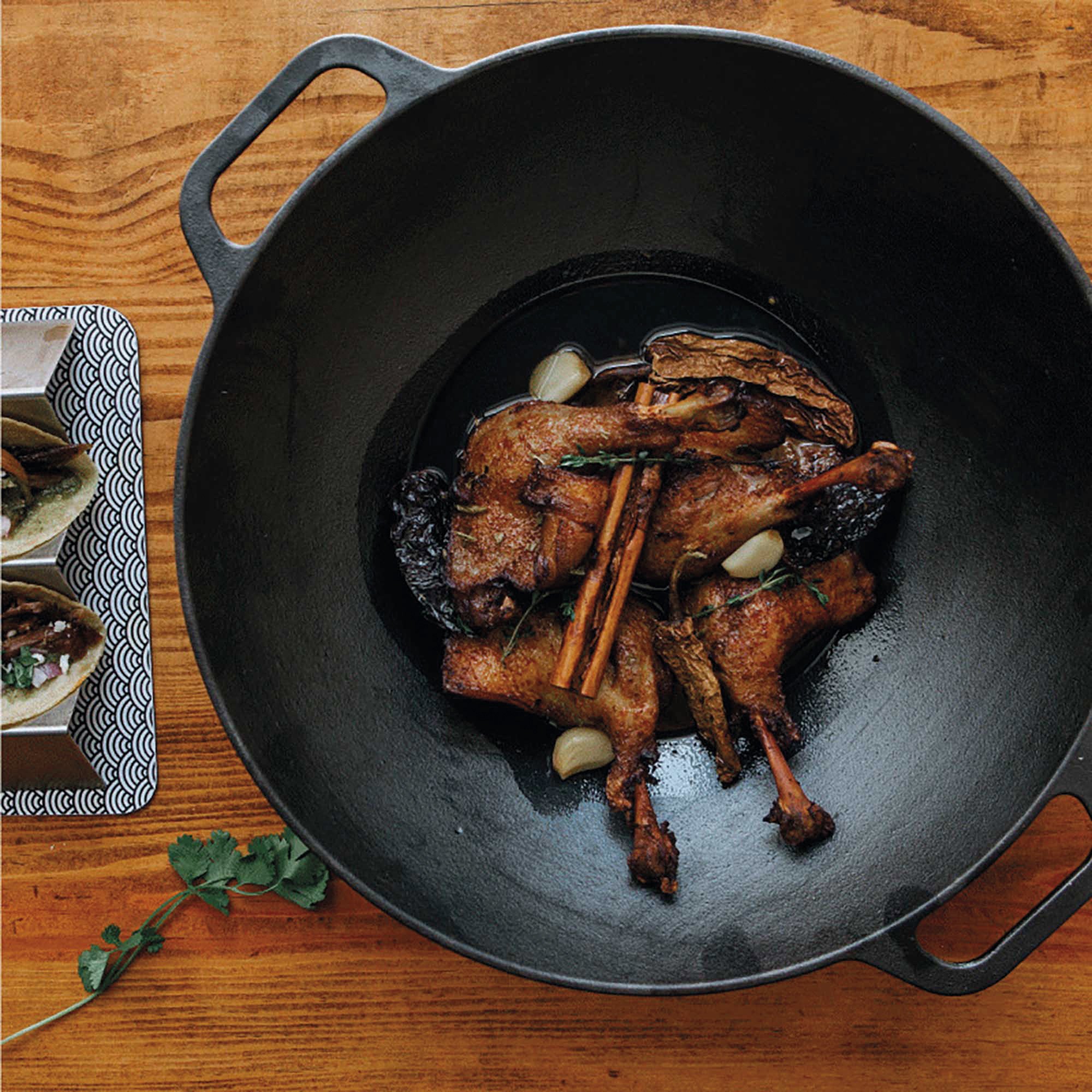 Enten-Confit in einer großen Pfanne mit Knoblauch und Gewürzen, neben Tacos auf Holz.
