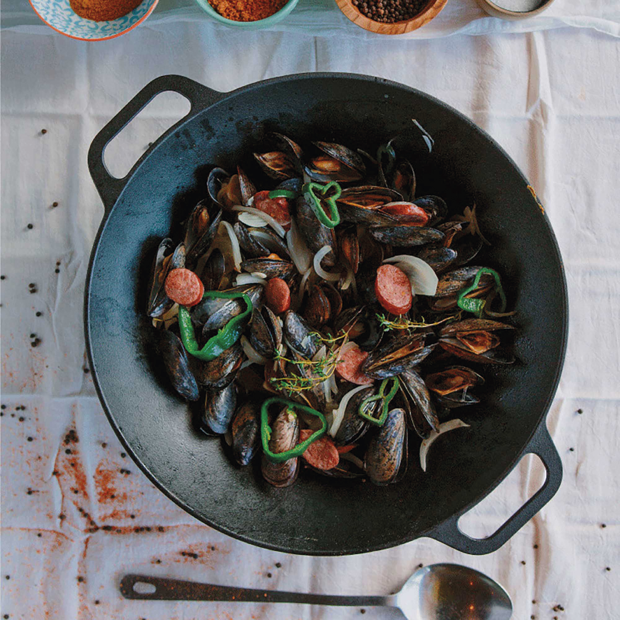 Muscheln in einer schwarzen Pfanne mit Würstchen, Zwiebeln und grünen Paprika. Weißes Tischtuch und Pfeffer.