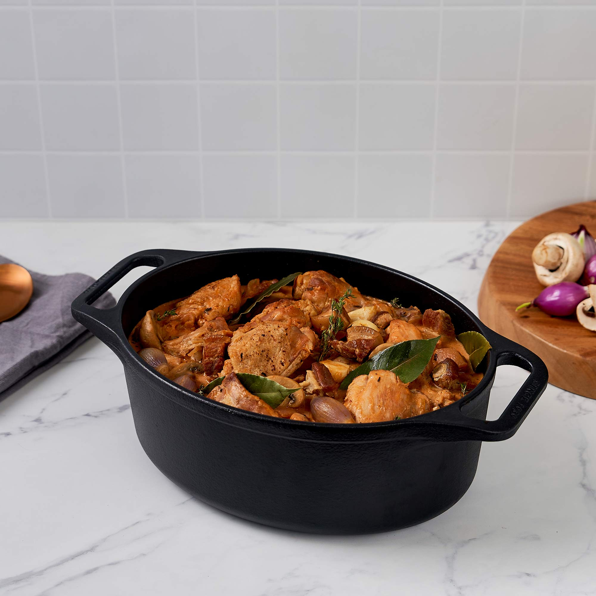 Una olla ovalada negra con estofado. Los ingredientes incluyen pollo, hierbas y cebollas. Fondo de mármol.