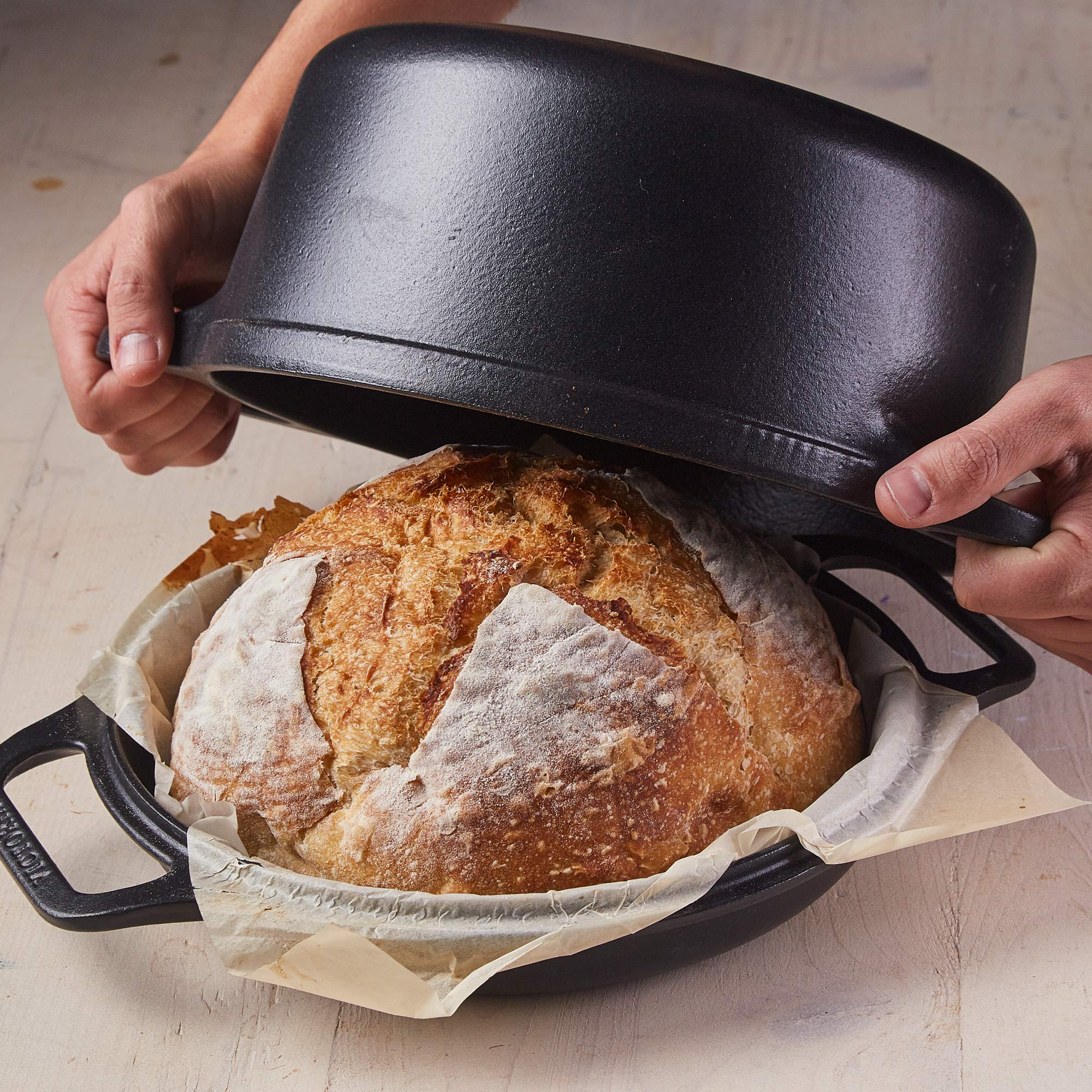 Manos levantan tapa negra de horno holandés, revelando pan horneado en papel pergamino.