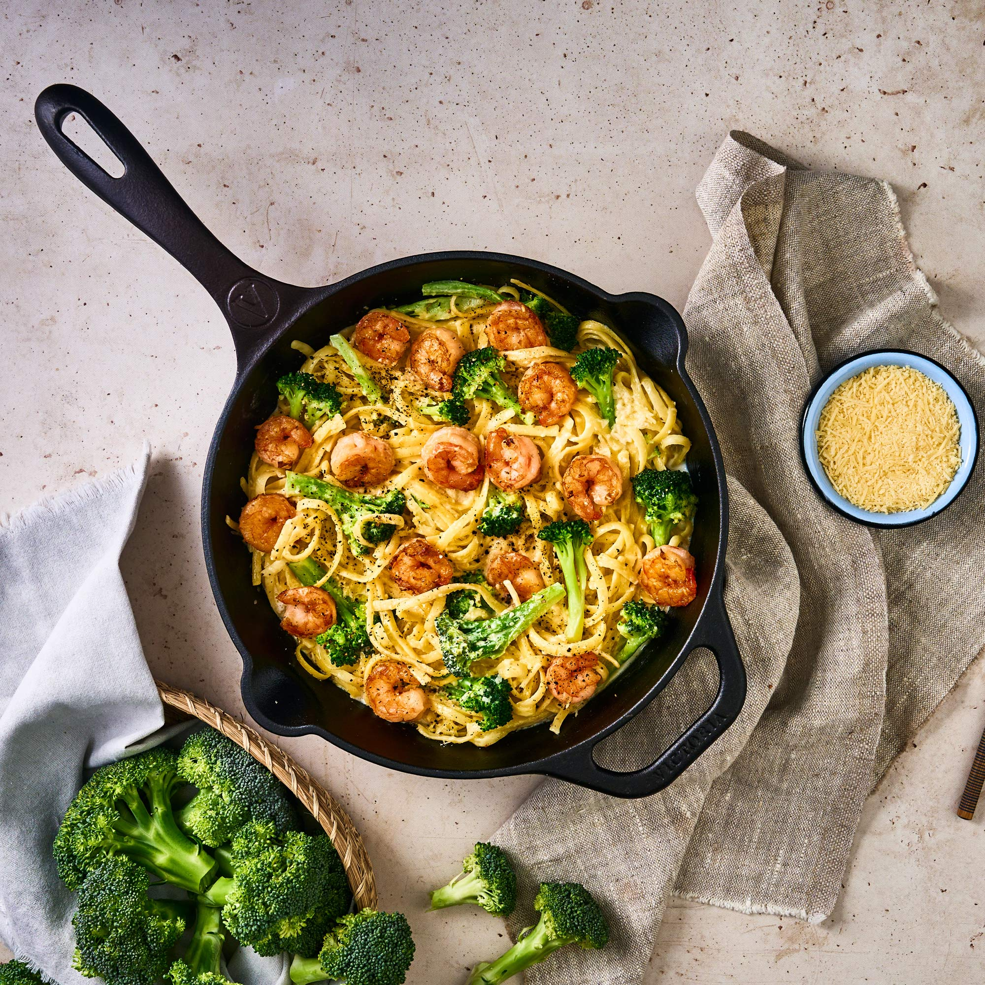 Sartén de pasta con camarones y brócoli, más queso rallado. Brócoli y tela en el fondo.