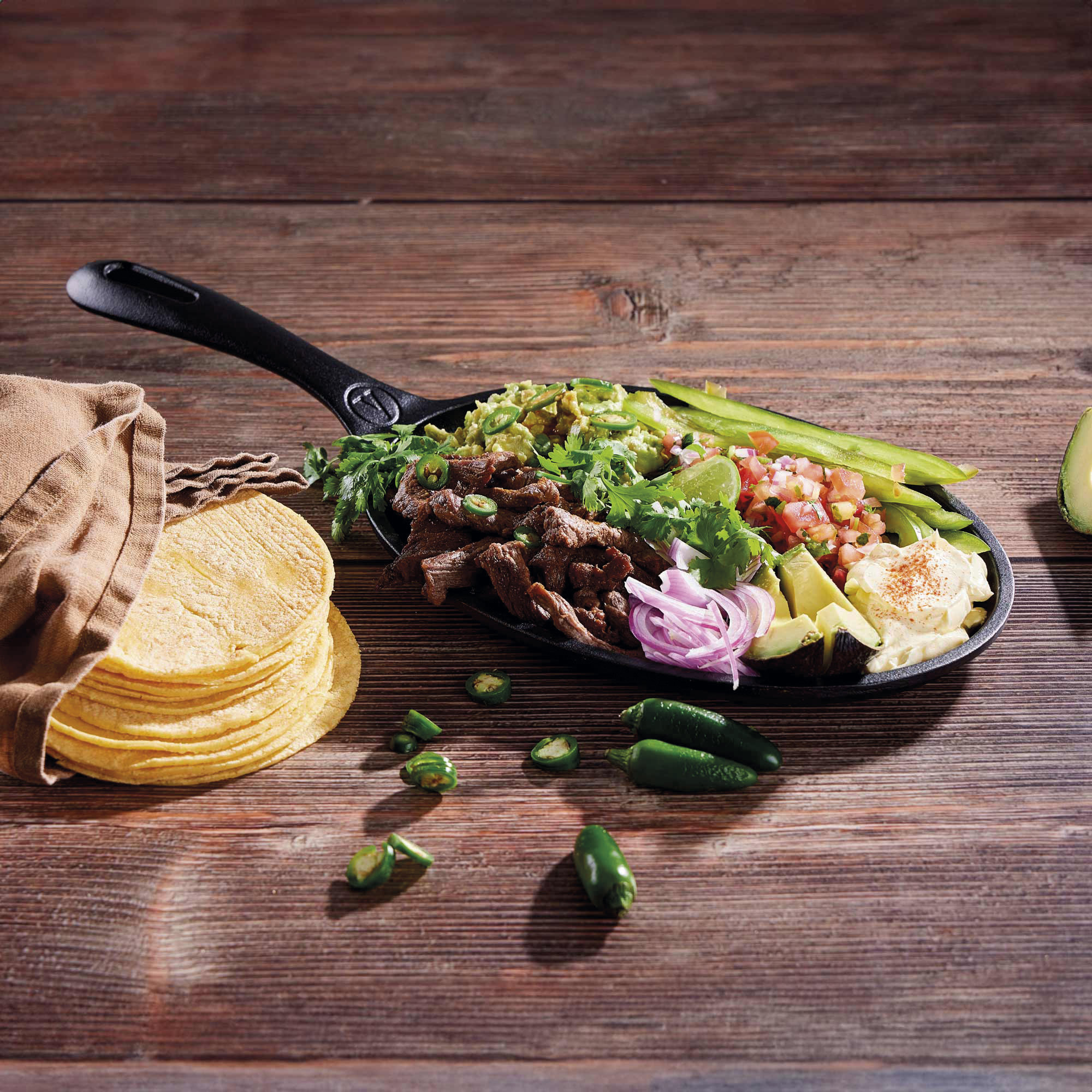 Eine Bratpfanne mit verschiedenen Zutaten. Enthält Tortillas, Fleisch, Guacamole, Zwiebeln, Jalapeños und Limette auf Holztisch.