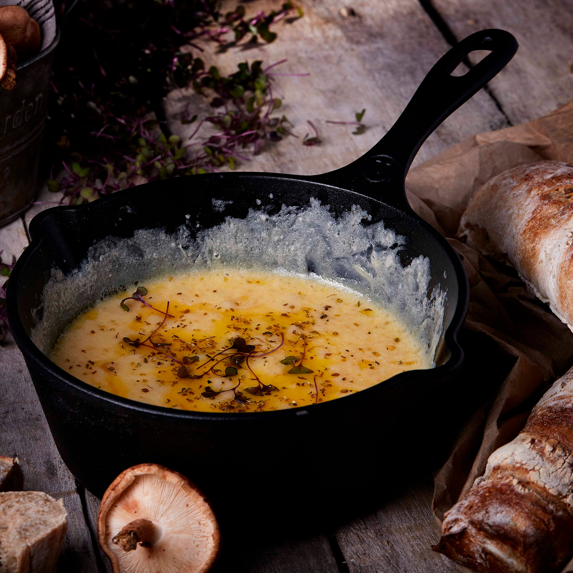 Una sartén de hierro fundido con queso derretido y hierbas. Pan y champiñones al lado.
