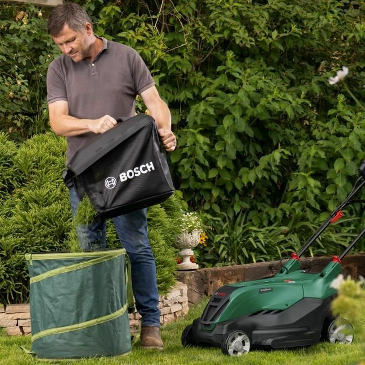 Hombre vaciando una bolsa negra en un contenedor verde junto a un cortacésped verde y negro en un jardín.