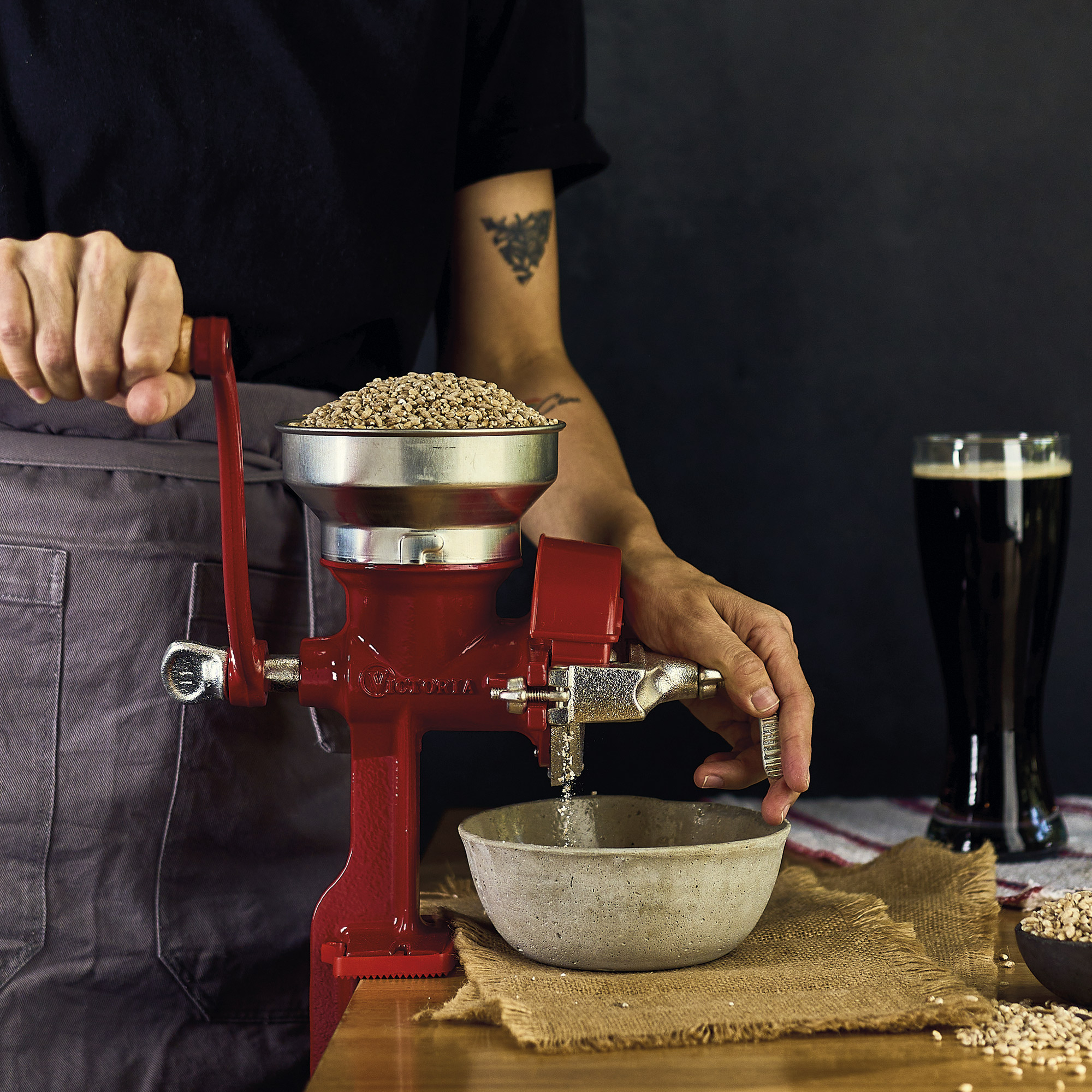 Eine Person verwendet eine rote Getreidemühle, um Getreide zu mahlen. Eine Schüssel und ein Bierglas stehen auf dem Tisch.